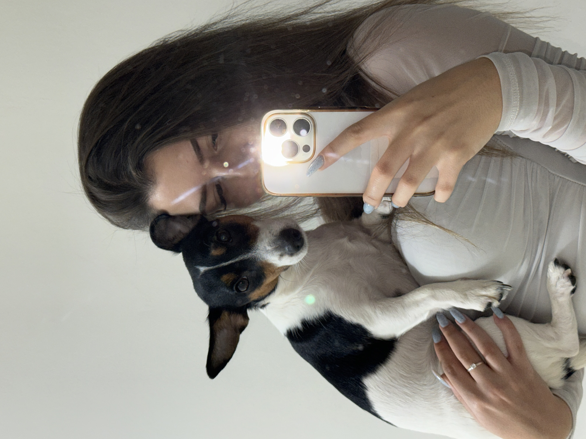 A woman is taking a selfie while holding a Jack Russell Terrier dog. The woman has long brown hair, light skin, and is wearing a sheer, light-colored long-sleeved shirt. She is holding a white smartphone with a gold frame; the flash is on, creating a glare. The dog is black and white, with its head near the woman's face. The background is a plain, light grey-white color. There is no evidence that suggests this image was taken in Sofia, Bulgaria.