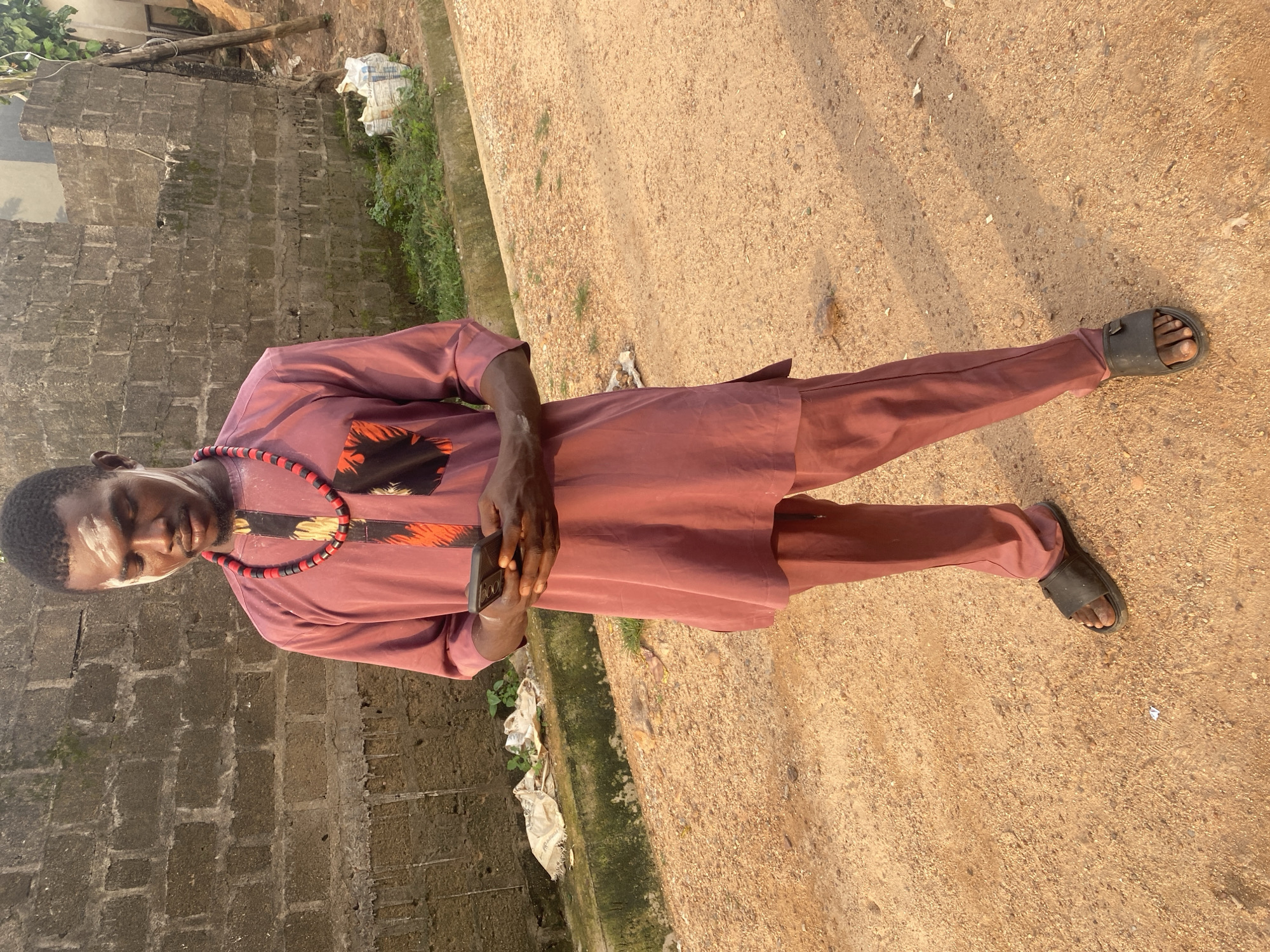 A man wearing a maroon-colored, long-sleeved tunic and matching trousers is standing on a dirt road in Afikpo, Nigeria. He is wearing black sandals and a beaded necklace. He is holding a black smartphone in his hands. To his left is a gray brick wall. Between the wall and the dirt road is an area of grass and weeds. There is trash on the ground near the wall. The dirt road extends into the background. The light is bright, suggesting it is daytime.