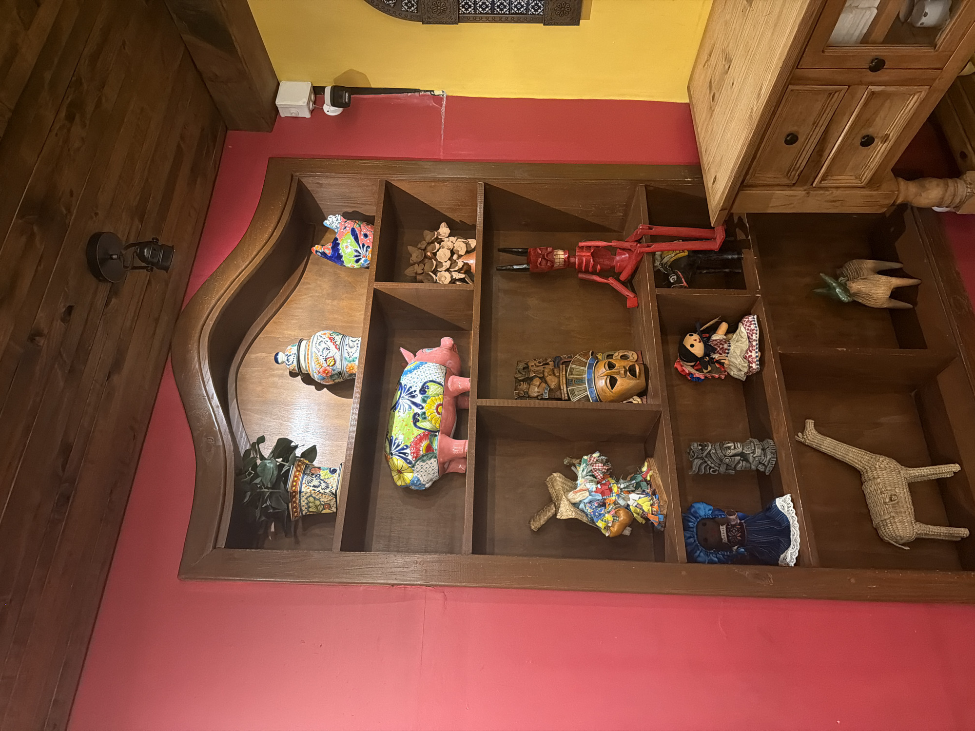 This image shows a wooden wall-mounted display case filled with a variety of decorative objects. The case is divided into several compartments, each containing different items such as ceramic pottery, wooden figurines, dolls, and plants. The case is mounted on a pink wall with a yellow section above. To the left of the case is a wooden wall with a black wall-mounted light fixture. To the right of the case is a wooden cabinet. The location is Unknown, Unknown.