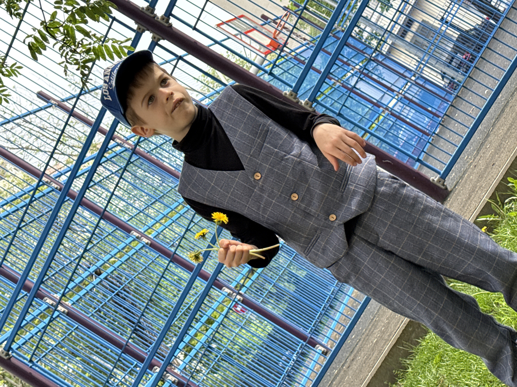 A young boy in a dark turtleneck, grey plaid vest, grey plaid pants, and a blue baseball cap stands outdoors, holding three dandelions in his left hand. The boy is standing next to a blue metal structure with horizontal and vertical bars. Behind the structure, a basketball hoop is visible. The location appears to be Moscow, Russia, based on the architecture and foliage visible. The boy is looking slightly upwards and to the right.