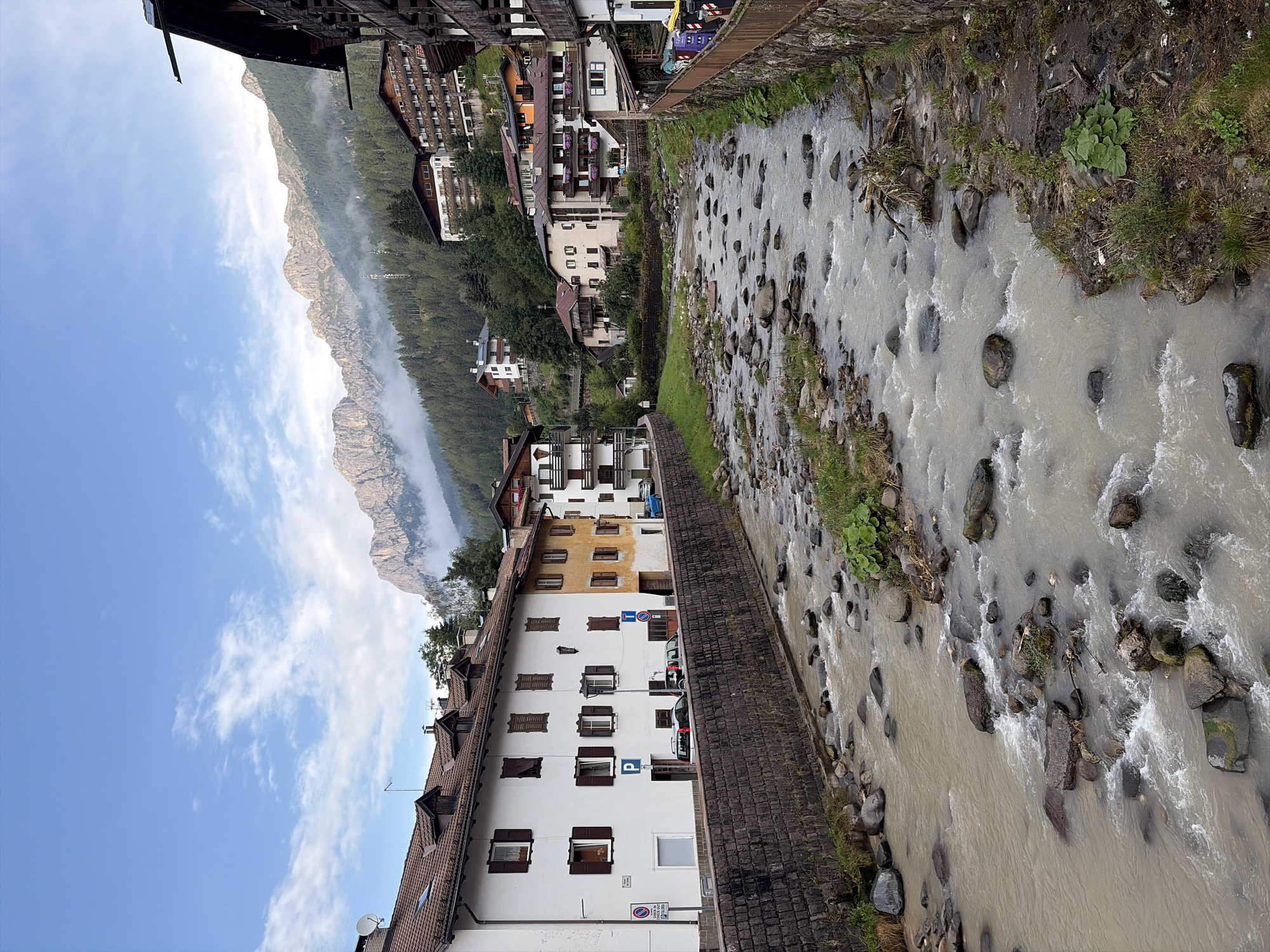 The image depicts a scenic view of the San Pellegrino stream flowing through the town of Moena, Italy. The stream, with its rocky bed and murky water, is flanked by stone walls and green banks. On the left, traditional alpine buildings with white and yellow facades, featuring dark wooden shutters and tiled roofs, line the street. Parking signs and cars are visible along the street. On the right bank, a wooden fence separates the stream from the town's buildings, which are characterized by wooden balconies and multi-story structures. In the background, lush green hills covered with pine trees rise steeply, leading to majestic, rugged mountains partially shrouded in mist under a blue sky with scattered white clouds. The overall scene conveys a tranquil, mountainous village atmosphere.