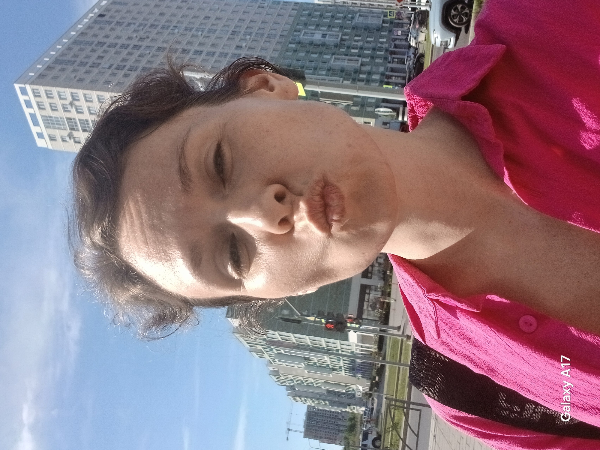 A woman poses in a selfie with an apparent "kissing" face in an urban environment. She wears a bright pink shirt, and a portion of a black garment is visible on her shoulder. The background features modern, high-rise buildings under a clear, blue sky, suggesting daytime. A sidewalk and street with cars are also present. Based on architectural features visible in the background, including distinctive high-rise buildings, the location is likely in Yekaterinburg, Russia. A watermark "Galaxy A17" is present in the lower right corner of the image.
