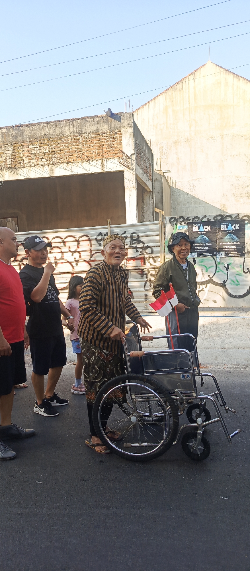 In Sekarpuro, Indonesia, a group of four individuals, including an elderly man in a wheelchair, stand on a street. The man in the wheelchair wears a striped outfit and a cap, while an Indonesian flag is attached to the wheelchair's handles. To his right, another man in a jacket holds a smaller Indonesian flag. To the left of the man in the wheelchair, two other men are visible, one in a red shirt and another in a black t-shirt. A young girl in a pink shirt and blue shorts stands between them, holding hands with one of the men. The street appears to be under construction, with a brick wall and fencing in the background, and graffiti on the fencing. A poster advertising "Djarum Black" is visible on the fence. The sky is clear and blue.