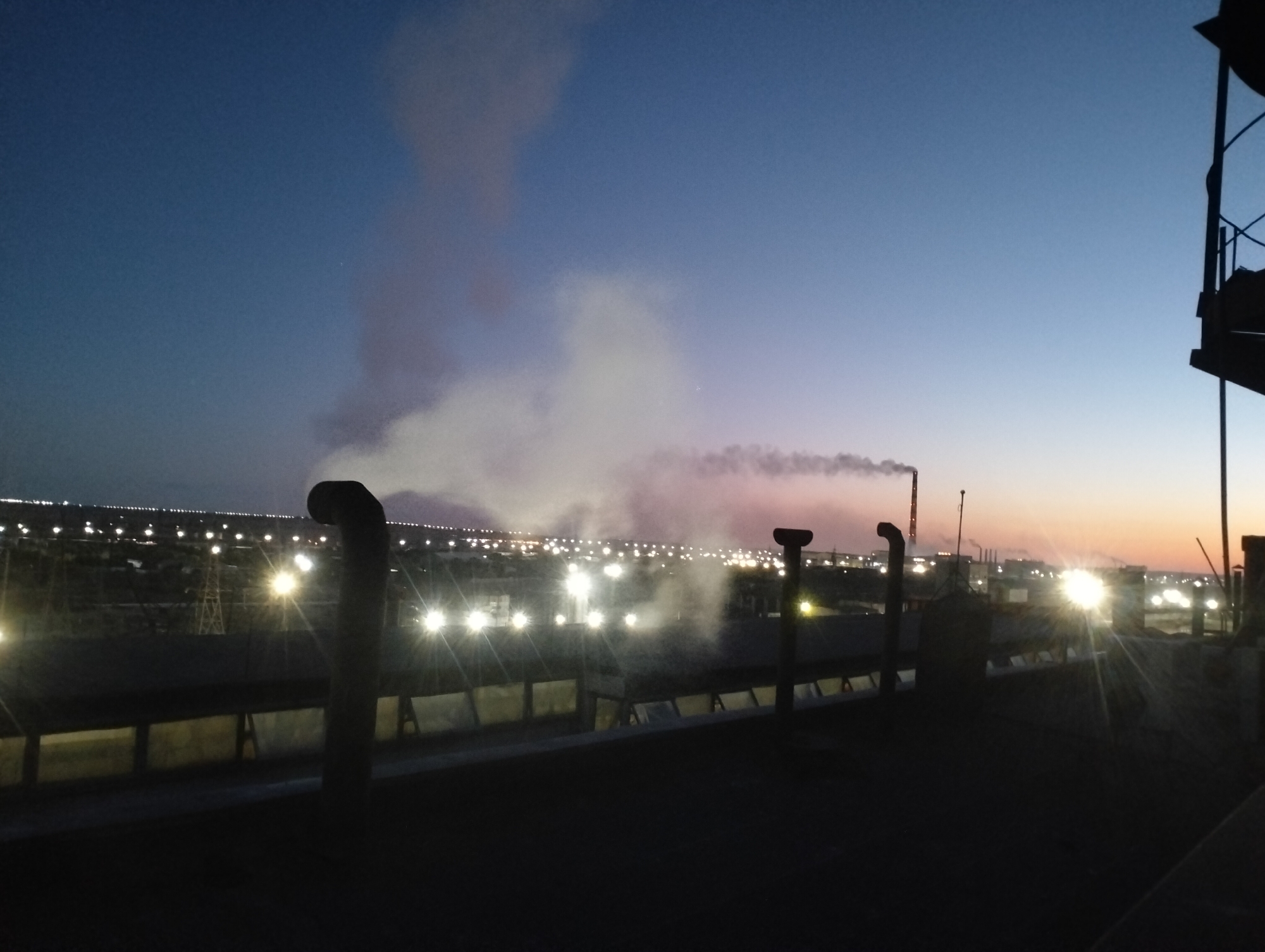 The image captures an industrial landscape at dusk in Jezqazğan, Kazakhstan. In the foreground, a dark rooftop area is visible, with several large pipes extending upwards. Beyond the rooftop, a sprawling city with numerous lights stretches across the horizon. The sky is a gradient of deep blue at the top, transitioning to lighter shades of blue and a faint pink or orange hue near the horizon, indicating the sunset or sunrise. Wisps of smoke or steam rise from various points in the distance, suggesting industrial activity. One prominent smokestack in the mid-distance is emitting a trail of dark smoke. The overall scene conveys a sense of industrial presence and the transition from day to night in an urban environment.