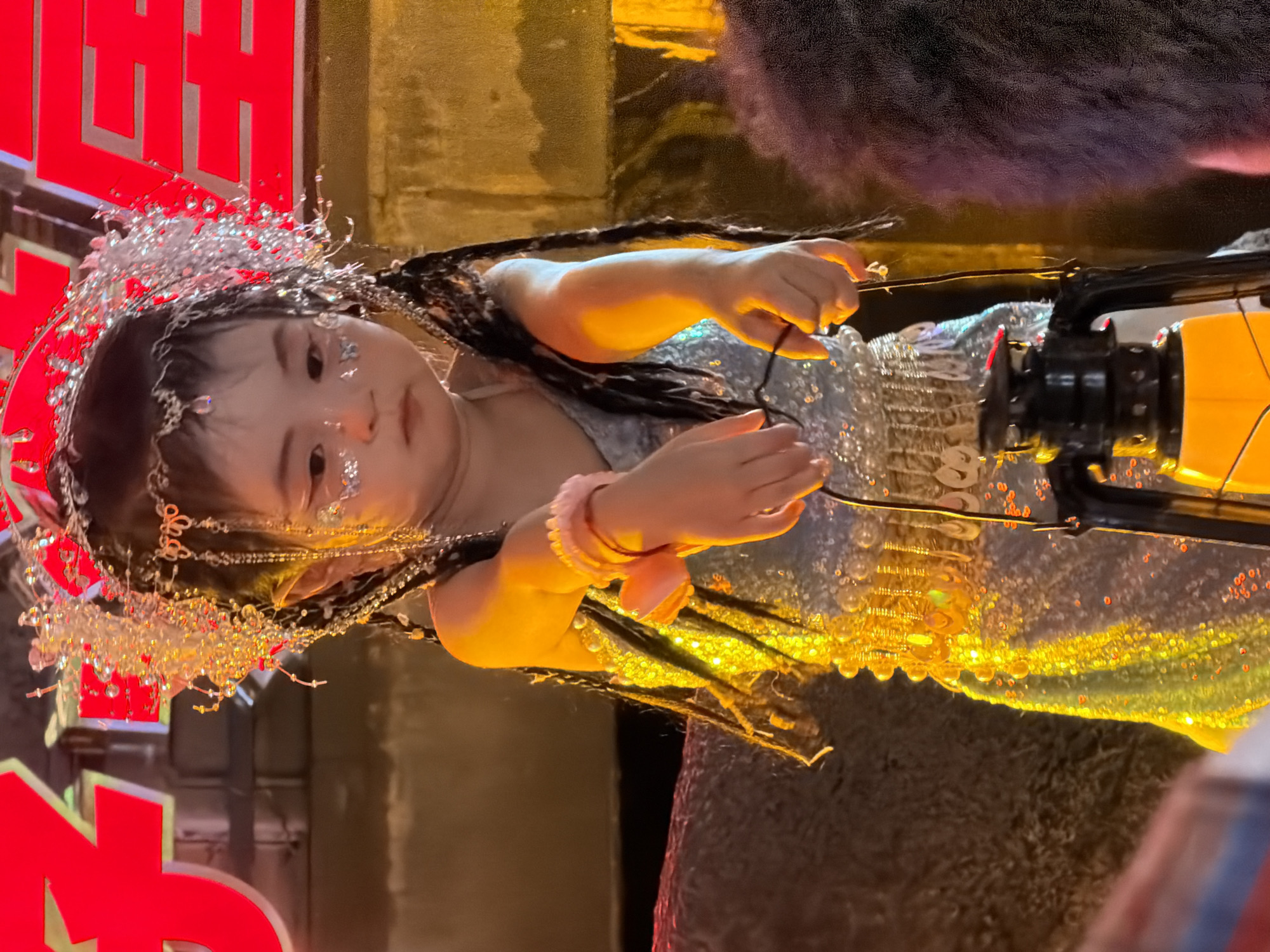 A young East Asian child, dressed in an elaborate, shimmering costume adorned with beads, sequins, and a jeweled headdress, stands holding a vintage lantern. The child's face is lightly painted with glitter, and their hair is styled in long braids. The background features a large, bright red neon sign with Chinese characters, suggesting a festive or commercial setting. The lighting is warm and yellow, casting a golden hue on the child and the surrounding environment. The image appears to have been taken at night, possibly during a festival or event, in the Yuzhong District of China, a well-known area for its vibrant urban landscape and cultural festivities.