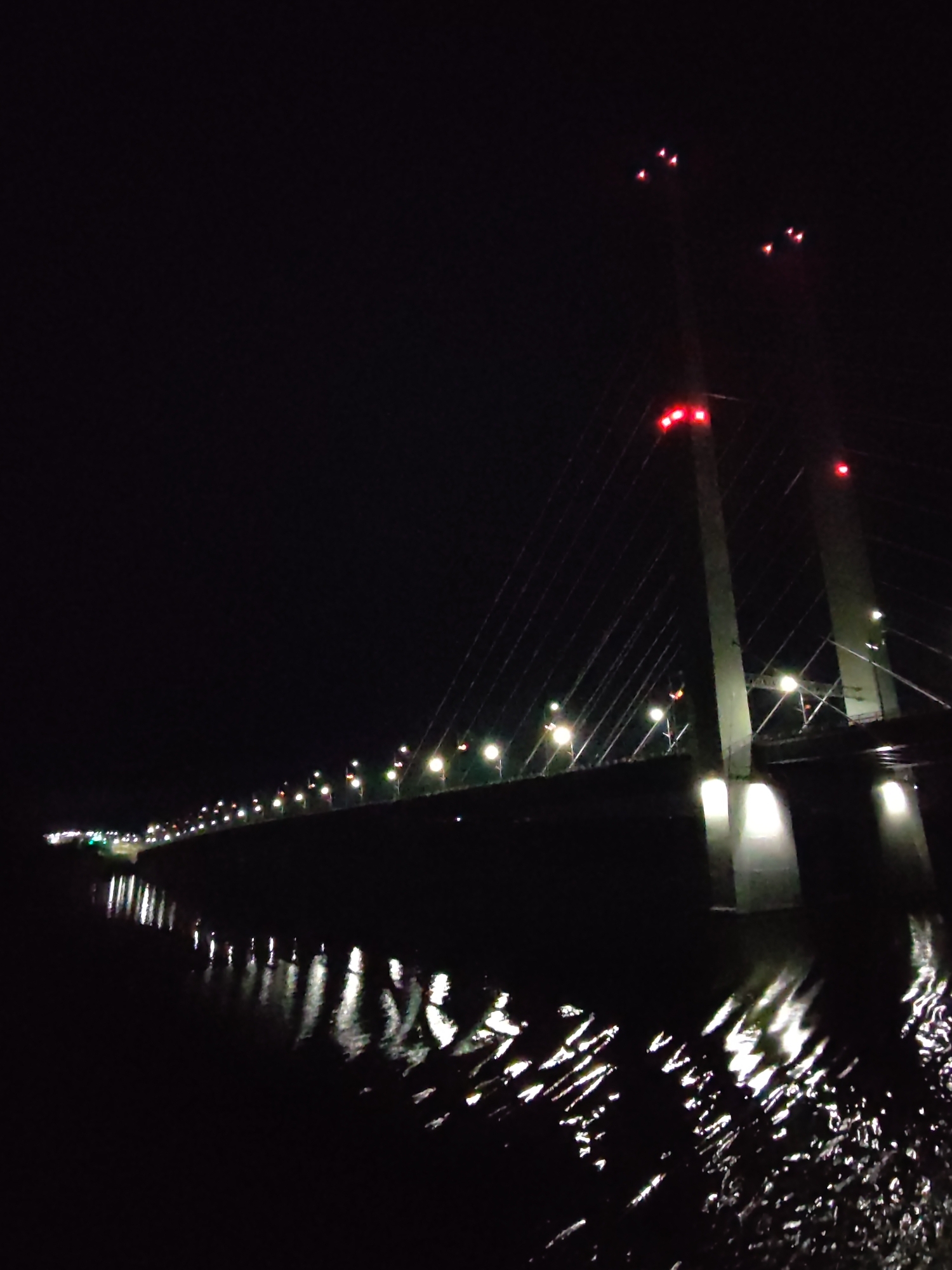 The image depicts the October Bridge in Cherepovets, Russia, at night. The bridge is illuminated with lights running along its length and red lights at the top of its two towers. These lights are reflected in the water below, creating streaks of white and red in the dark water. The overall scene is dark, with the bridge and its reflections providing the main sources of illumination.