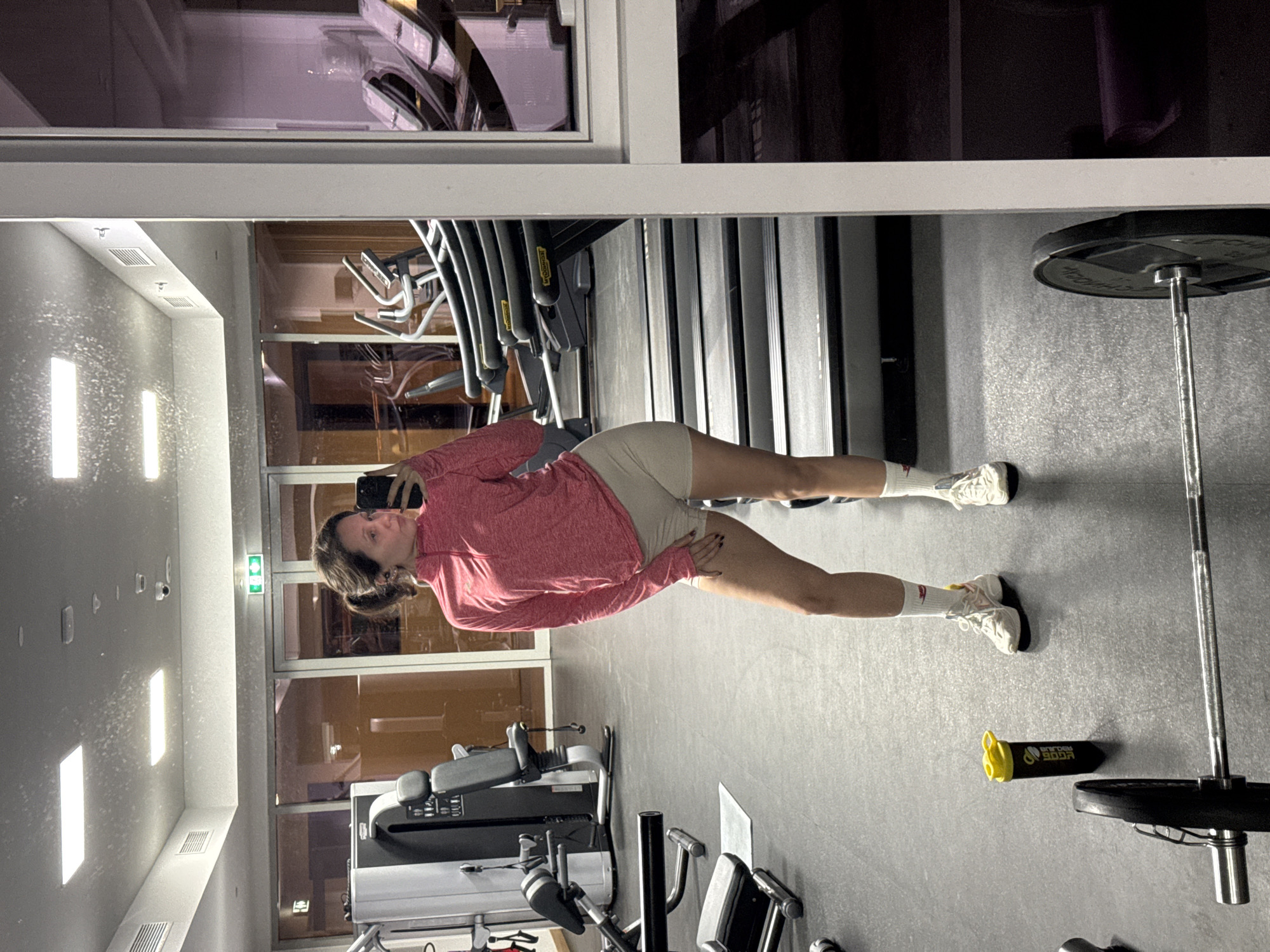 The image depicts a woman in a gym, taking a selfie in front of a mirror. She's wearing a pink long-sleeved shirt, beige shorts, white socks, and white sneakers. The gym contains treadmills, weightlifting equipment including a barbell with weights, and other exercise machines. The gym appears to be indoors, brightly lit with artificial lights on the ceiling. The reflection in the mirror shows details of the gym interior. Based on the equipment and modern design, the location is potentially in a commercial or residential fitness center. Without additional information, a precise location within Dubai cannot be determined.