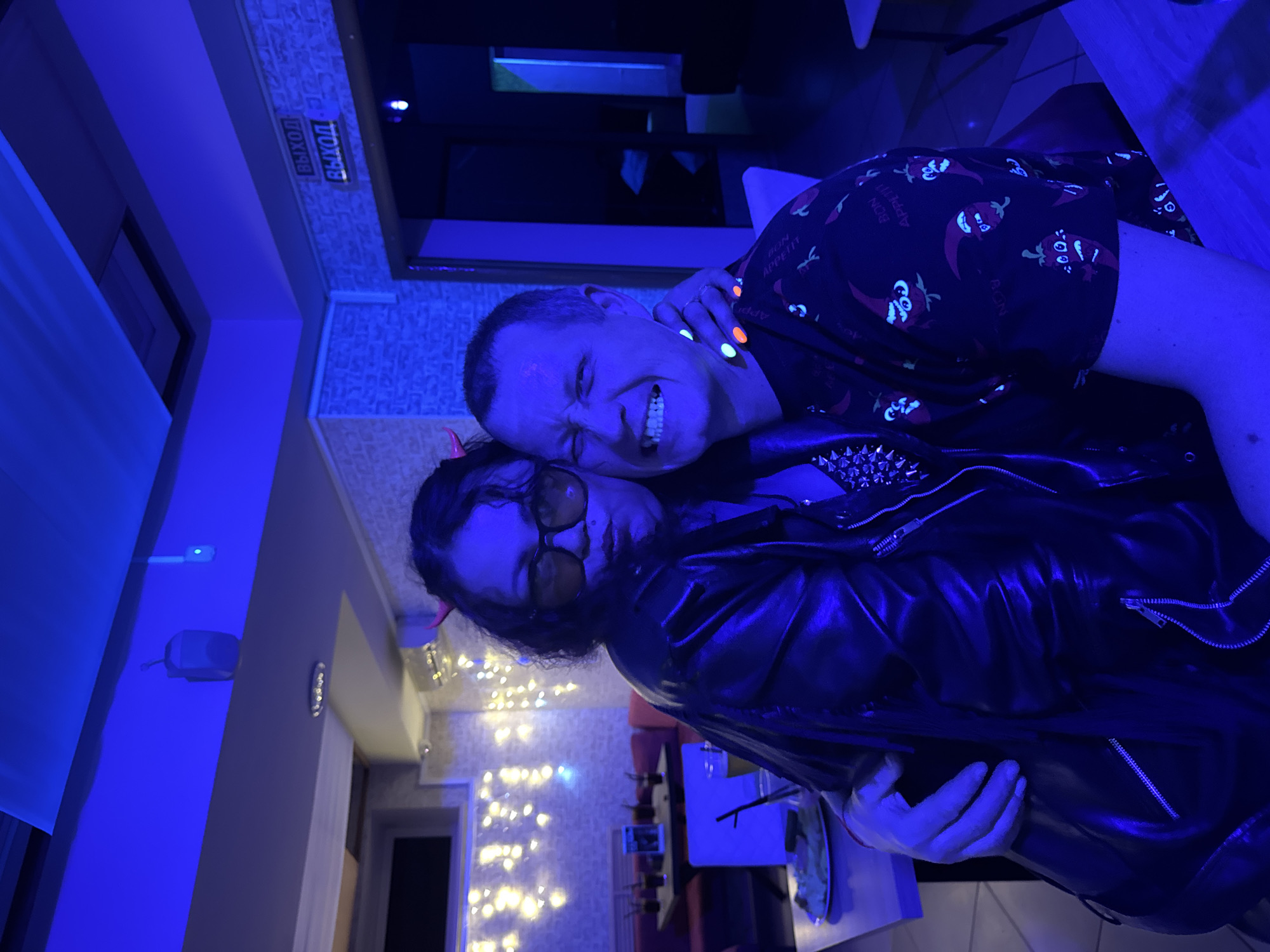 Two individuals are posing in an indoor setting, lit with predominantly blue lighting. The person in the foreground is wearing a black leather jacket with silver studs and orange-tinted glasses. The person behind them is wearing a black shirt with red pepper motifs and appears to have neon-colored accessories on their wrist and fingers. Both individuals appear to be smiling or grimacing for the photograph. In the background, there are string lights, a door, and signs written in Cyrillic. Based on information retrieved, the location is the city of Chaikovsky, Russia.