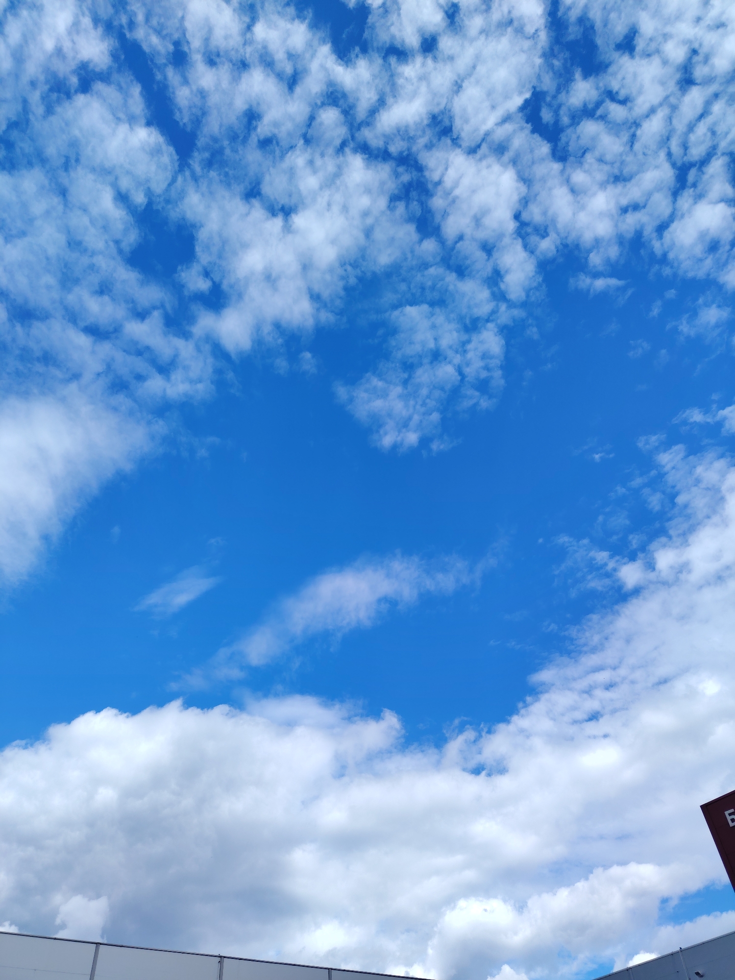 This is a photograph of a bright blue sky filled with scattered white cumulus clouds. The clouds are dense and fluffy, with some appearing wispy and stretched across the sky. In the foreground, a portion of a white building or structure is visible along the bottom edge of the image, with a dark red sign with white Cyrillic lettering on the right side. The sign appears to have the letter "Б" visible. The scene suggests an outdoor environment, possibly in a location like Хоругвино, Russia, given the visible text. There are no people or animals depicted in the image. The lighting indicates it is daytime.