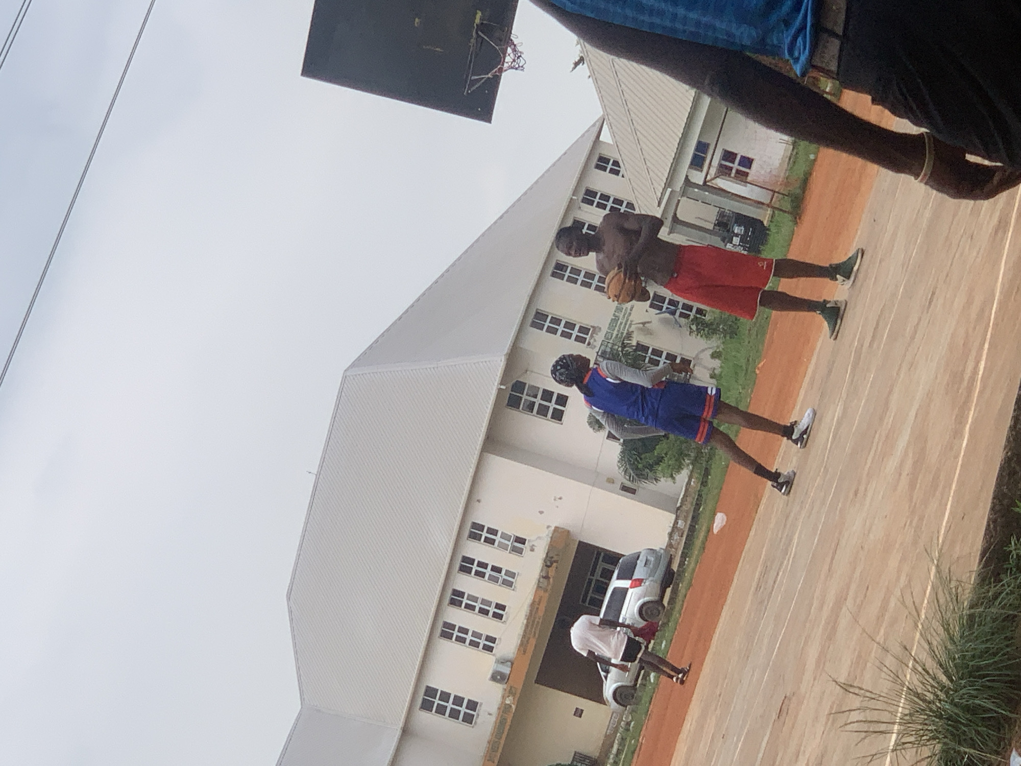 In Ukukwa, Nigeria, two young men are engaged in a basketball game on an outdoor court. One player, shirtless and wearing red shorts, holds a basketball, preparing to shoot or pass. He is positioned to the right of the frame. To his left, another player, dressed in a blue and orange basketball uniform, appears to be on defense, anticipating the next move. The court is made of a reddish-brown material, with white lines marking its boundaries.
Behind the court, a large, cream-colored building with a white, angled roof dominates the background. The building has numerous windows, suggesting it might be a school or administrative facility. A silver car is parked near the building, partially visible. The sky is overcast, indicating a cloudy day, and the lighting suggests it is daytime. The atmosphere seems casual and energetic, typical of a friendly game.
The image is rotated, with the subjects appearing on a diagonal. The overall scene depicts a community gathering or recreational activity taking place in an outdoor urban or semi-urban environment in Nigeria.