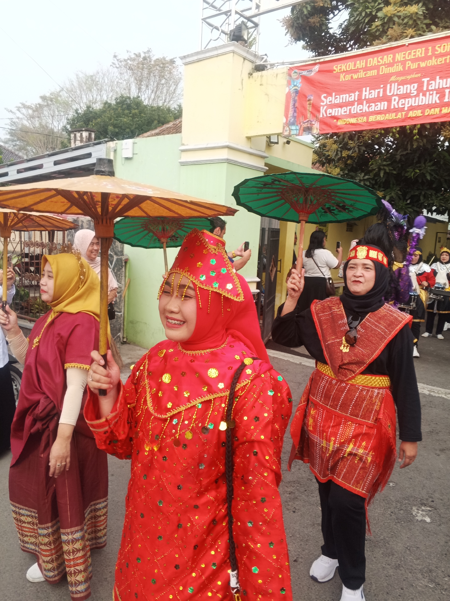 In Sokanegara, Indonesia, a group of women are participating in a parade, dressed in vibrant, traditional attire. The foreground is dominated by a young woman in a bright red, sequined costume and a matching headdress, holding a large yellow umbrella. To her left, another woman wears a maroon outfit with a gold hijab, also holding a yellow umbrella. Behind them, a woman in a black shirt and a red and gold woven skirt stands with an umbrella. In the background, a banner reads "SEKOLAH DASAR NEGERI 1 SOK..." and "SELAMAT HARI ULANG TAHUN KEMERDEKAAN REPUBLIK IN..." suggesting a celebration of Indonesian Independence Day. Other participants, including those in drum corps uniforms, are visible further down the street. The scene appears to be taking place on a paved road lined with buildings and trees under an overcast sky.