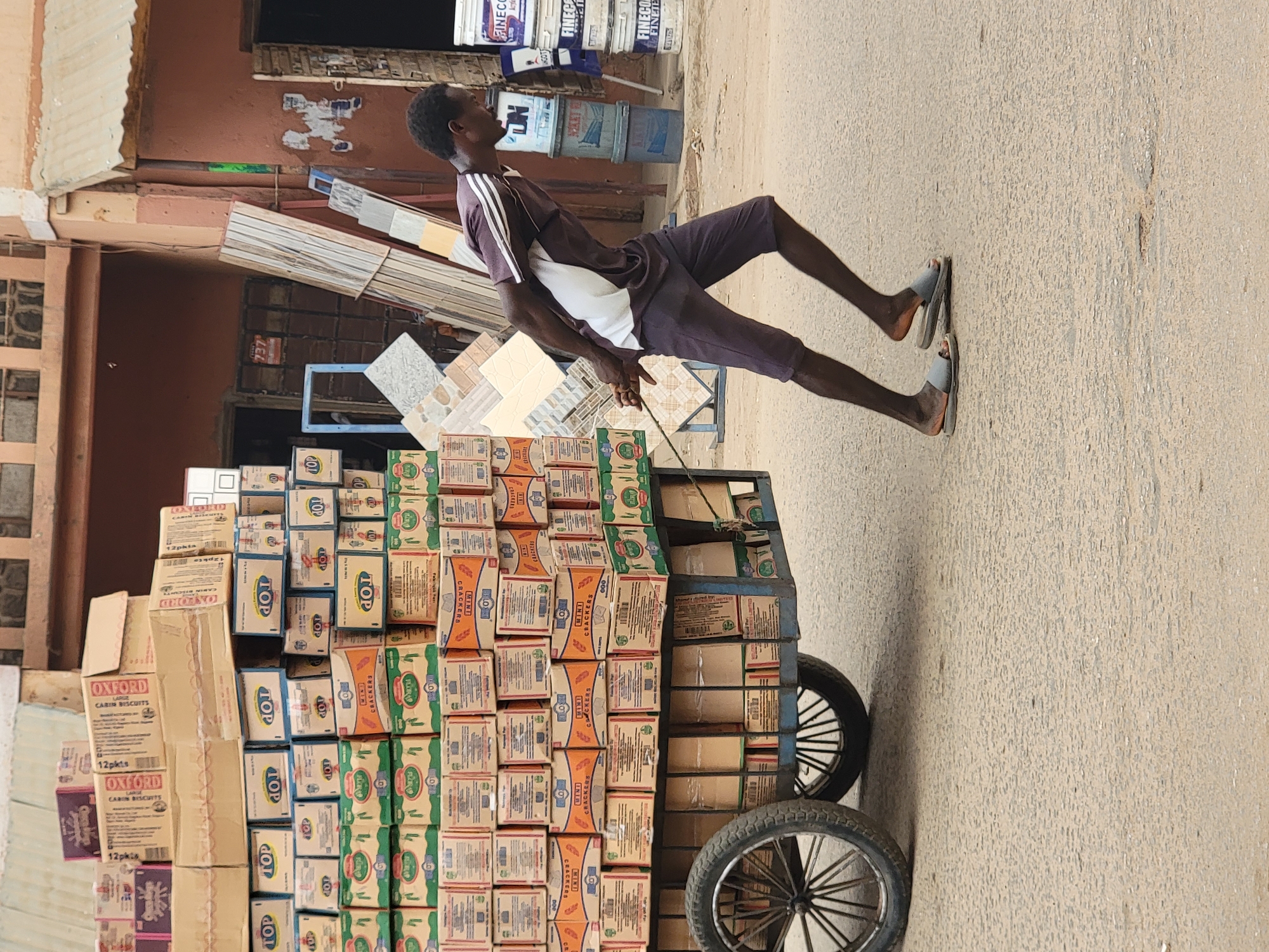 A man in Maiburiji, Nigeria, pulls a heavily loaded cart of biscuits down a dusty street. The cart is stacked high with boxes of various brands of biscuits including "OXFORD", "TOP", and "FLOUR", and has two wheels. The man wears a dark-colored athletic shirt with white stripes, dark shorts, and sandals. The environment includes buildings and street surfaces. The presence of construction materials suggests possible building work nearby.