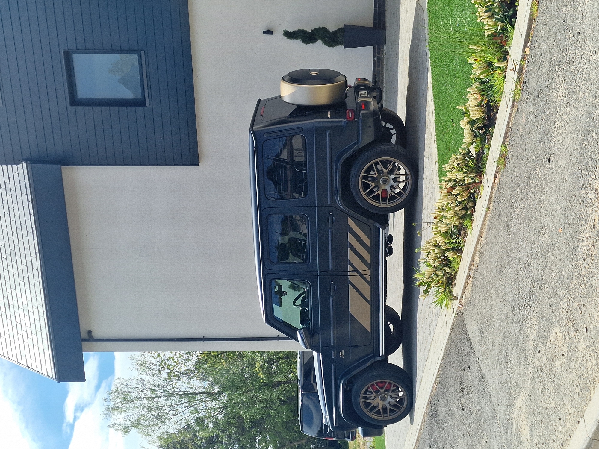 A black Mercedes-Benz G-Wagon with gold accents is parked on a paved driveway in front of a modern house. The house has a light-colored exterior wall and a dark blue vertical paneling section with a square window. There's a patch of green lawn between the driveway and the house, and a neatly trimmed evergreen topiary in a black pot stands next to the house. The location is Witton Gilbert, United Kingdom. The lighting suggests it's daytime.
