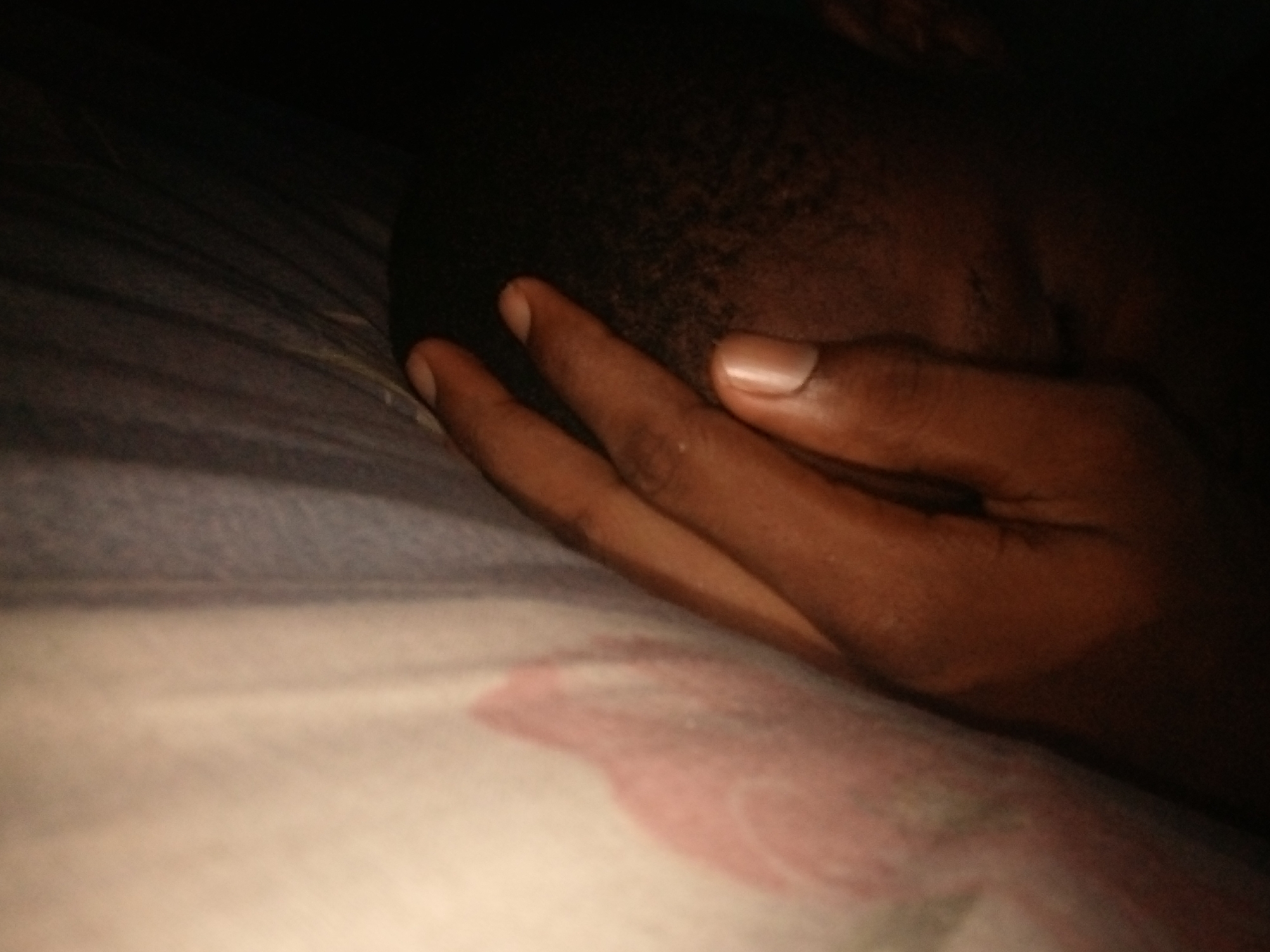 The image shows a close-up, dimly lit view of a person's head and hand resting on a bed with patterned sheets. The person's face is obscured by their hand, which is covering their eyes and forehead. The skin tone appears to be dark. The sheets have a pattern that includes light colors and a reddish-pink floral design. The background is dark, suggesting the image was taken in low light conditions. The scene appears to be indoors, likely in a bedroom. There are no visible elements to indicate a specific location within Shangisha, Nigeria.