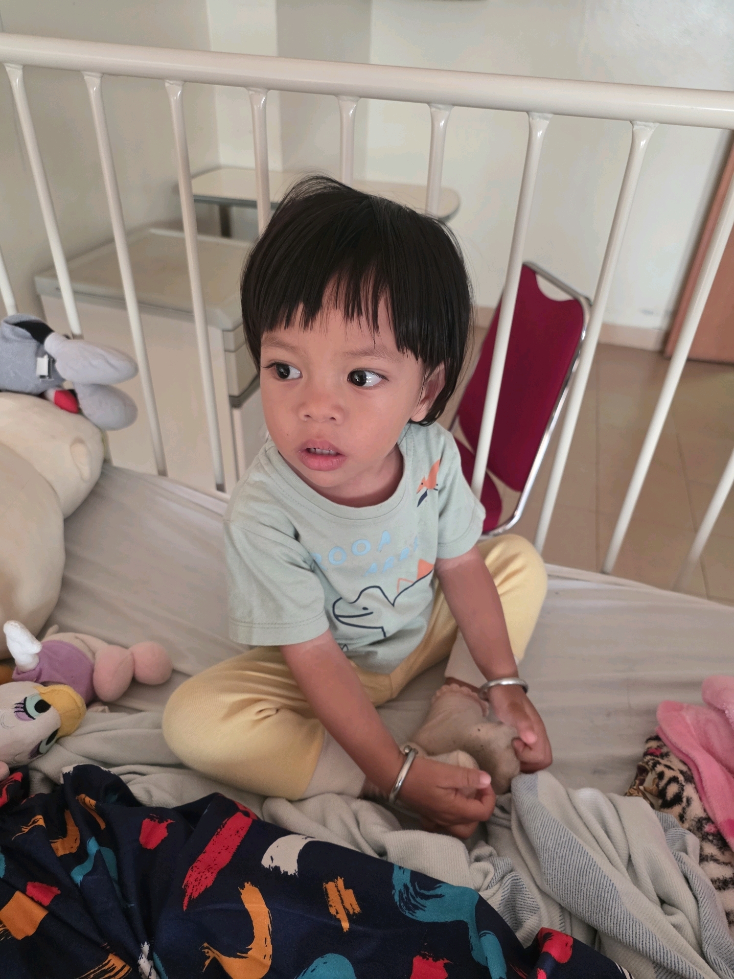 A young child with dark hair and fair skin sits in a white metal crib. The child is wearing a light green t-shirt with a dinosaur graphic and yellow pants. The child's hands are clasped together, and they are looking to the left with an inquisitive expression. In the crib, there are several stuffed animals, including a large white one, a purple and pink one, and a grey one with red accents. A patterned blanket with abstract brush strokes in red, orange, white, and blue is also visible in the crib. The background shows white walls and a red chair. The overall impression is that this is a medical setting, possibly a hospital room for children, within the city of Tanjung Pinang, Indonesia.