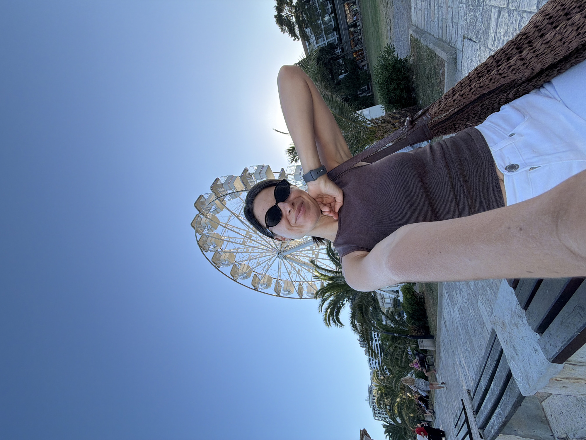 The image displays a woman taking a selfie on a paved promenade in Budva, Montenegro. The woman, positioned in the foreground and slightly to the right, is wearing a dark brown ribbed tank top, white jeans, and dark oval-framed sunglasses. A dark grey smartwatch is visible on her left wrist, and a dark brown woven bag is slung over her right shoulder. Her left arm is bent with her hand near her chin, and she exhibits a slight smile. Her right arm, extending to hold the camera, is partially visible at the bottom left.
The background features a wide, light-colored stone-paved walkway with several wooden benches supported by concrete bases on the left. Multiple indistinct figures are visible walking or sitting on these benches in the mid-ground. Numerous tall palm trees and dense green bushes punctuate the mid-ground. Behind the vegetation, multi-story buildings with multiple balconies, typical of coastal architecture, are discernible. Dominating the upper-middle and right background is a large white Ferris wheel with numerous gondolas, extending high into a clear, bright blue sky. A bright light source, consistent with the sun, is visible behind palm trees to the right of the Ferris wheel.