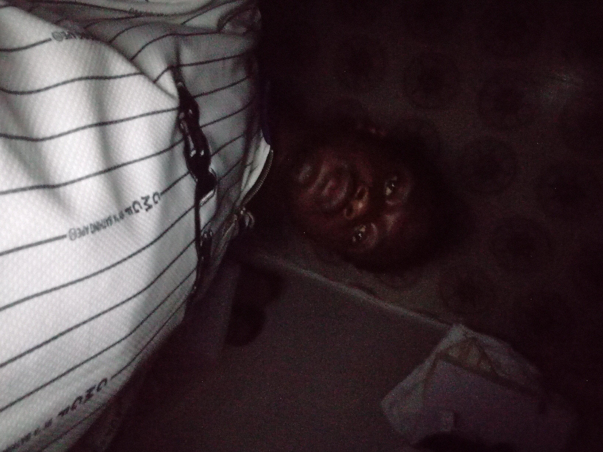 This dimly lit image, potentially taken indoors in Ogbogbo, Nigeria, features a person's face and a striped bag. The lighting is very poor, suggesting it was captured at night or in a room with minimal illumination.
The primary subject is a person with dark skin, whose face appears in the lower-right portion of the frame. Their head is tilted, and they are looking directly upwards towards the camera with open eyes. Due to the severe darkness, their expression is difficult to discern, but they appear awake.
To the left, a white fabric item with prominent horizontal black stripes dominates a large section of the image. This appears to be a bag, possibly a travel bag or backpack, as indicated by a visible black zipper running vertically along its right edge. Upon closer inspection, several words are faintly visible on the striped fabric. "OWO" can be seen near the top of the bag, and "OZOR" is visible upside down towards the bottom. Other smaller, less legible text may also be present, possibly related to a brand or item description.
The background is uniformly dark, appearing to be a floor or a solid surface, possibly with a subtle pattern in some areas. The camera perspective is unusual, suggesting a high angle looking down, or the subject might be lying down. There is no discernible activity or specific event taking place beyond the subject looking at the camera. The overall scene is casual and candid, characterized by its low light and intimate framing.