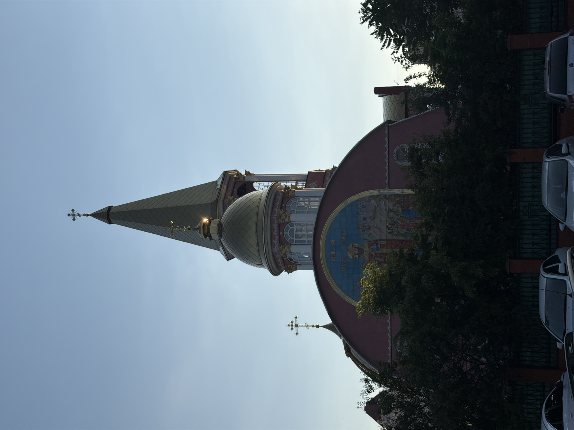 The image depicts the Church of the Ascension of the Lord in Kyiv, Ukraine, at dusk. The sky is a clear, pale blue, with a hint of sunset. The church's dominant feature is its steep, dark spire, topped with a gold cross. Below the spire sits a golden dome, also adorned with a cross. The church's exterior is painted a reddish-pink and features a large, arched mosaic depicting a religious scene. Trees with dark green foliage obscure the lower half of the church. In the foreground, a green metal fence runs horizontally, and several parked cars are visible. The lighting suggests it is late afternoon or early evening.