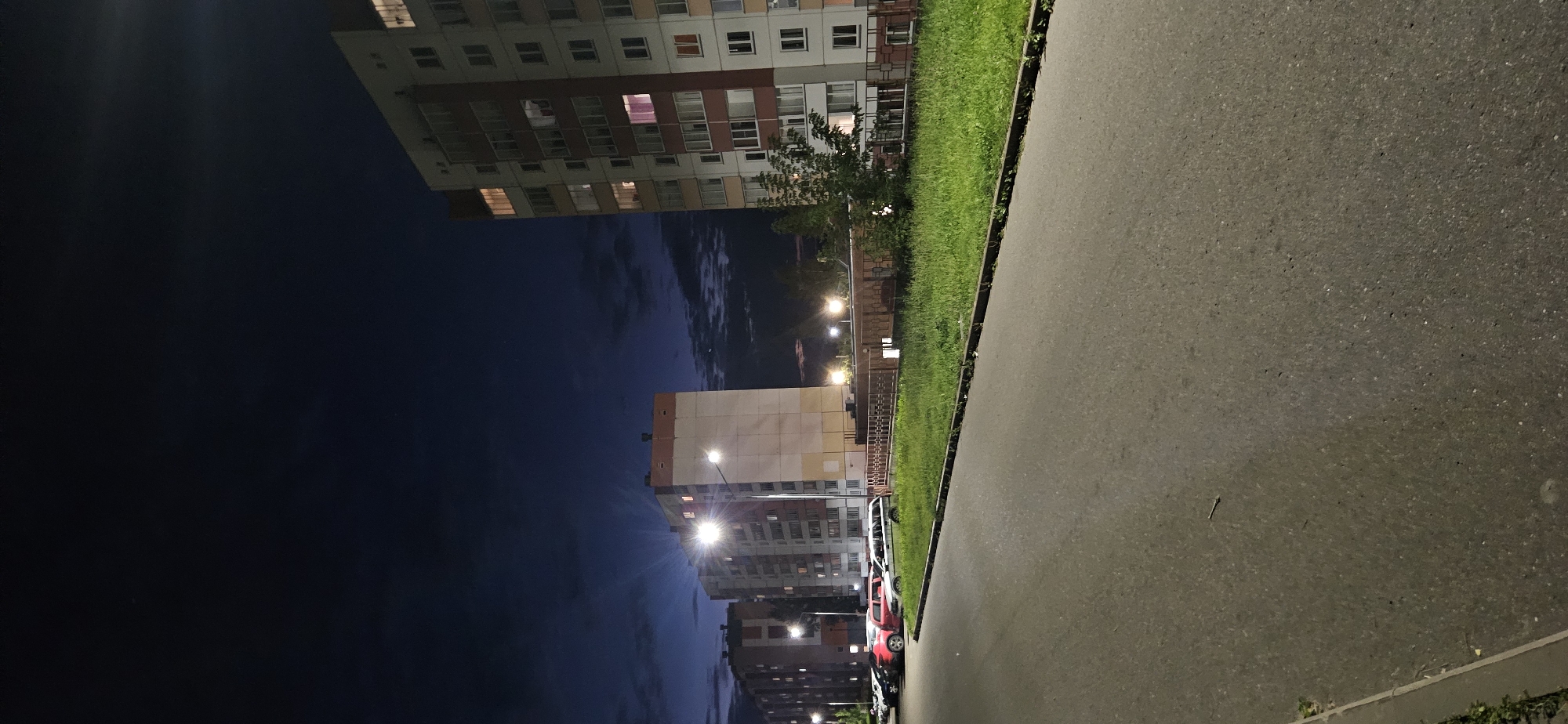 An outdoor, vertical, medium shot captures a nighttime scene on a paved road in a residential area, likely in Vitsebsk, Belarus. The sky is a deep, dark blue with scattered wispy clouds, and the only light sources are streetlights and illuminated apartment windows. Two tall, multi-story apartment buildings dominate the right and center-left of the frame. The building on the right is predominantly white with red and orange accents, featuring numerous windows, some of which emit a warm, orange glow, while others have a pinkish hue. The building on the left is a lighter beige with a pale orange band near the top, also displaying illuminated windows. A patch of green grass runs along the side of the road in front of the buildings, bordered by a low, reddish-brown fence in the middle ground. Several cars are parked on the left side of the road, including a red SUV, a white station wagon, and other vehicles obscured by darkness and distance. A single streetlight stands near the center of the frame, casting a bright light upwards and outwards. The paved road occupies the foreground, showing signs of wear and a light-colored curb on the left edge. A single cigarette butt is visible on the road. The overall atmosphere is calm and quiet, characteristic of a residential neighborhood at night.