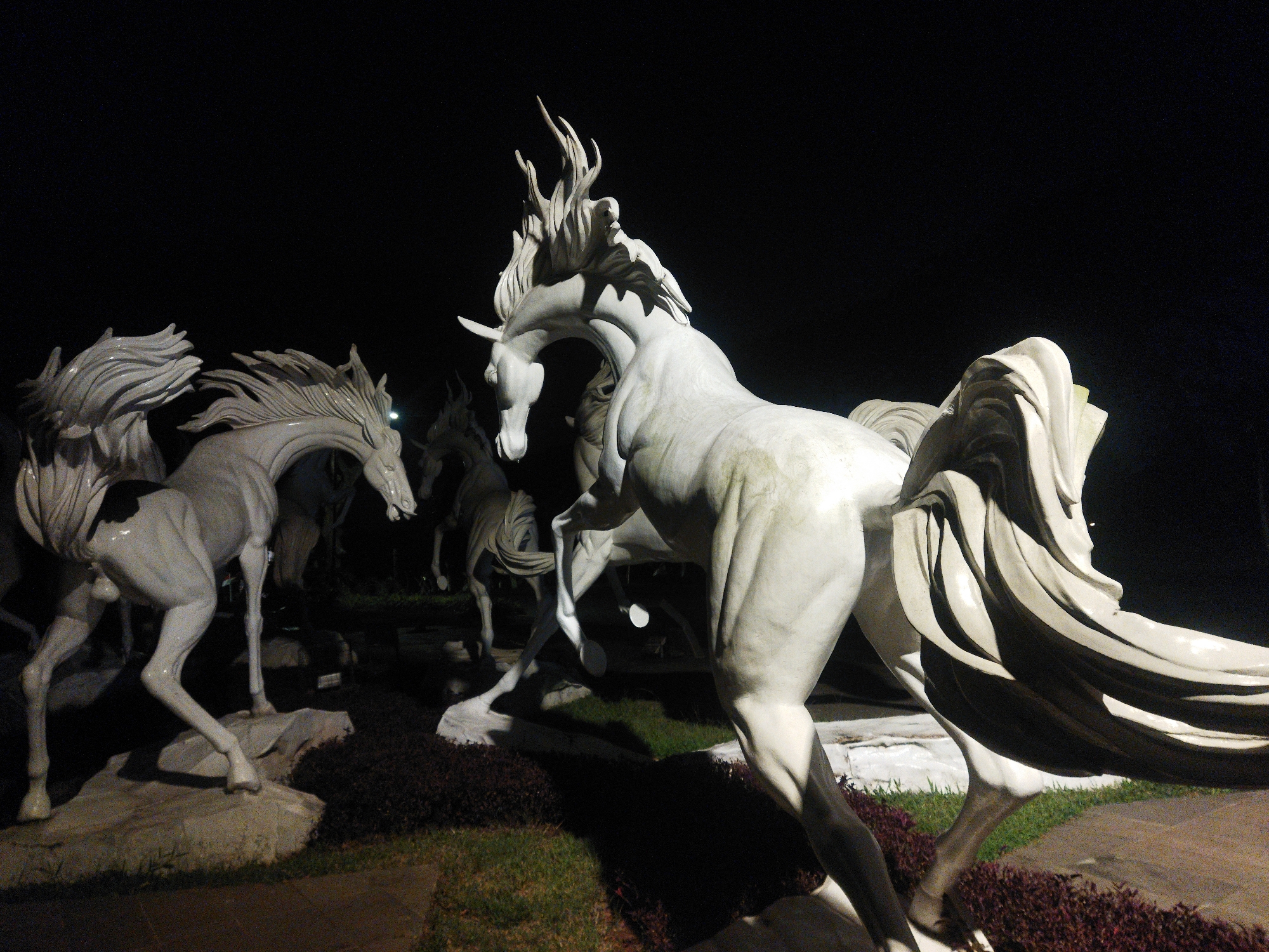 This is a nighttime photograph of several white horse sculptures in a landscaped area. The sculptures appear to be made of marble or a similar stone and are dramatically lit, casting long shadows. The horse in the foreground is rearing up, with its mane flowing behind it, while other horses are depicted in various dynamic poses. The background is dark, suggesting it is night. The sculptures are situated on rocky terrain with patches of grass and dark red, low-lying plants. The overall impression is one of powerful movement and artistic expression.
The image depicts a section of the Sam Poo Kong Temple complex in Semarang, Indonesia. Specifically, this sculpture garden is located within the temple grounds. The Sam Poo Kong Temple is a Chinese temple dedicated to Admiral Zheng He, a Chinese explorer. The sculptures are a modern addition to the historical site, designed to represent the spirit of exploration and strength, often associated with horses in Chinese culture.