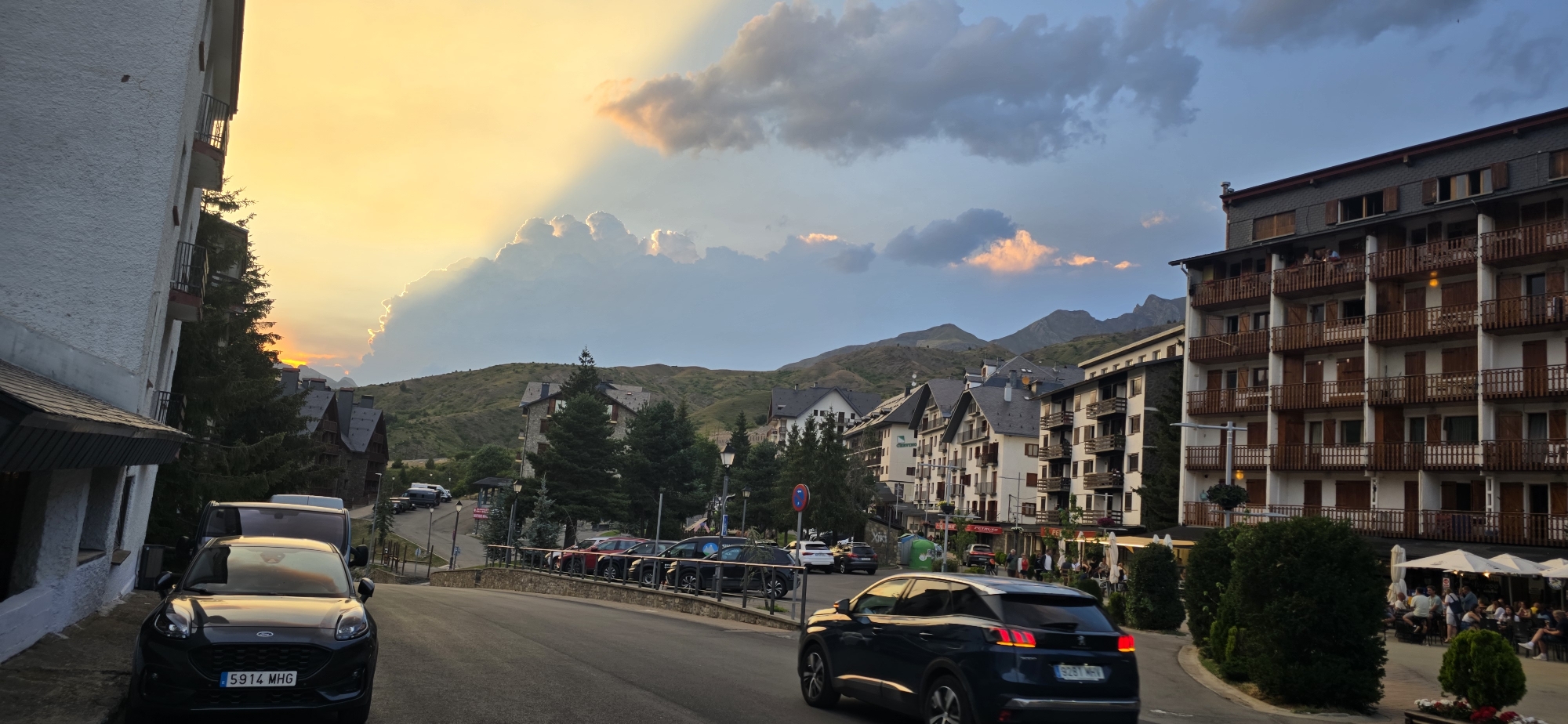 The image shows a street scene in Sallent de Gállego, Spain, during what appears to be sunset or late afternoon. The sky is dramatic with a blend of warm golden light and muted blue, pierced by rays of light from behind a large bank of clouds. In the foreground, a dark SUV is parked on the left side of the road, and another dark SUV is driving away from the camera on the right side of the road. The road curves uphill, lined with buildings of varying heights and architectural styles, characteristic of a mountain town. Many of the buildings have balconies with wooden railings.
To the left, a portion of a white building with a textured facade is visible. Beyond it, traditional-looking buildings with dark pitched roofs are nestled amongst evergreen trees. Further up the street, larger apartment buildings with multiple floors and numerous balconies are prominent. Some of these buildings have shops or restaurants on the ground floor, with outdoor seating areas and umbrellas visible. In the distance, rolling hills and mountains form the backdrop. The scene appears to be a street in a resort town, possibly with a ski resort nearby given the architecture. The lighting suggests a pleasant evening with some residual warmth from the setting sun.