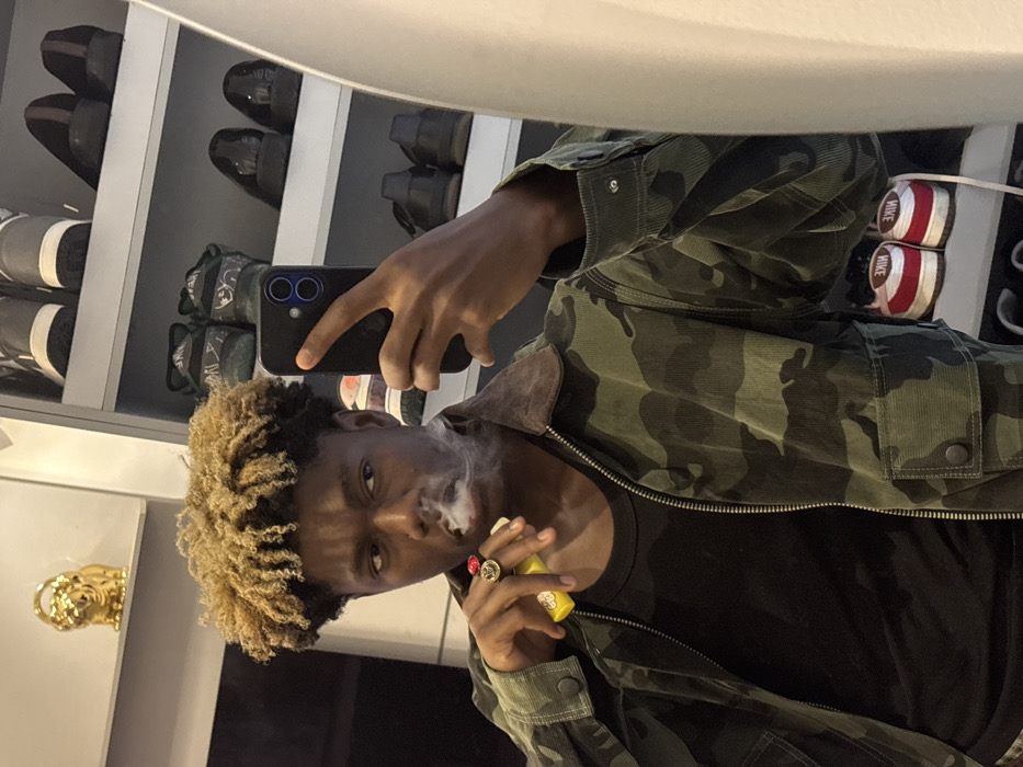 An indoor scene depicts a young adult male with dark skin and bleached blonde, curly hair taking a mirror selfie. He is wearing a black t-shirt beneath an unzipped camouflage-patterned corduroy jacket with a brown collar. A ring with a red top and gold-toned base is visible on his left ring finger. He holds a black smartphone with a dual-lens camera in his right hand, angled to capture his reflection. In his left hand, he holds a yellow vaping device, from which he exhales a cloud of white vapor. His gaze is directed slightly off-camera to his right. The background features a white shelving unit stocked with various pairs of shoes, including dark formal shoes, dark athletic shoes, and a pair of red and white Nike sneakers. A gold-colored figurine, resembling a lion, rests on a shelf above the subject's head. The specific location in Igboefon, Nigeria, cannot be determined from the visual evidence.