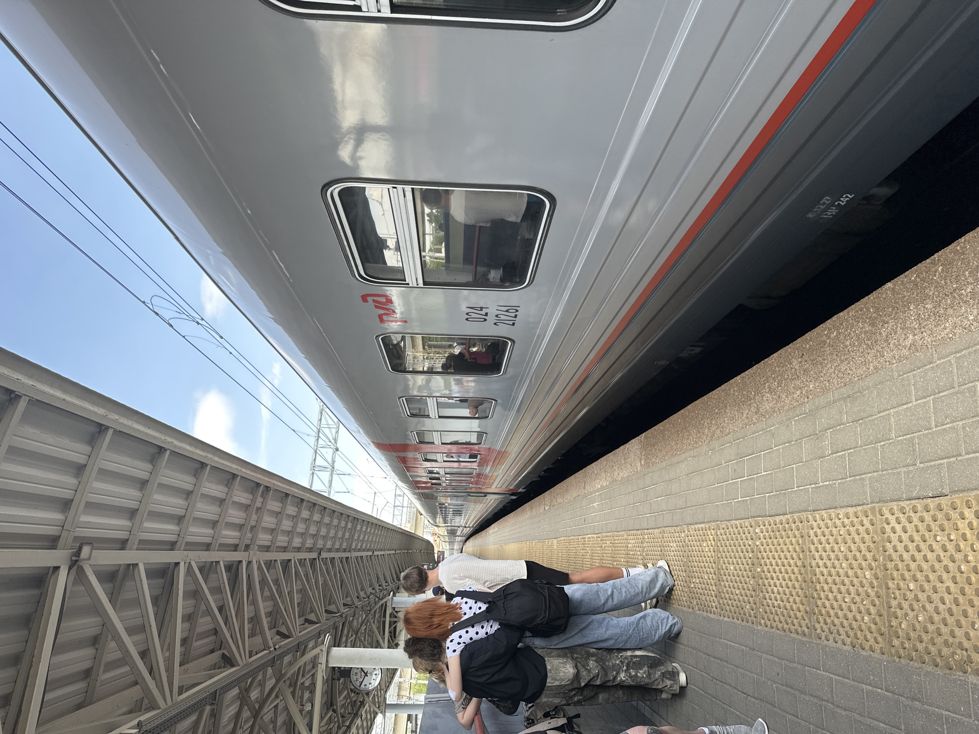 The image shows a train station platform in Nizhny Novgorod, Russia. A gray train is stopped alongside the platform. A group of people stand on the platform, appearing to wait for the train. The platform is paved with yellow textured tiles near the edge, and gray rectangular tiles further back. Above the platform, there is a gray metal roof structure. Electrical wires run above the train. The train has the "RZD" logo and the number "024 21261" printed on its side.