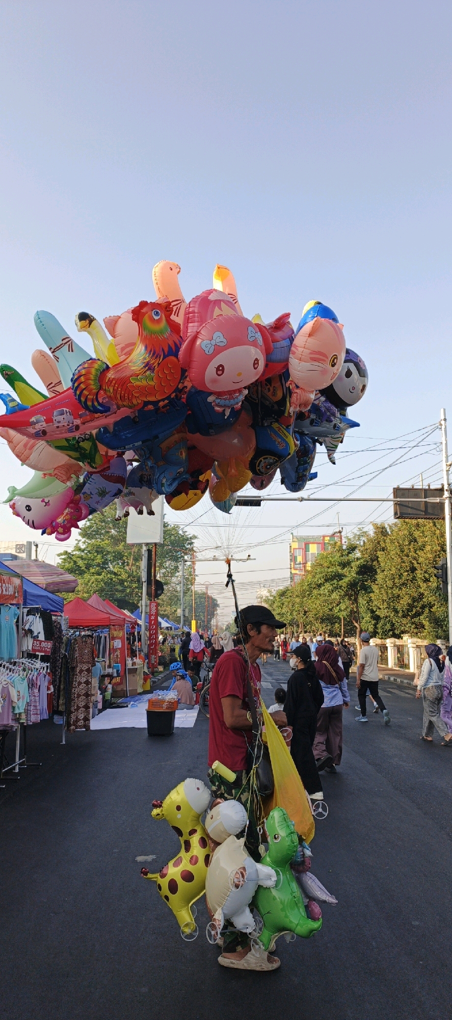 The image depicts a street scene in Sidoarjo, Indonesia, likely a market or roadside gathering, captured during daytime with clear sky. A man in a red shirt and black cap stands in the foreground, holding a large bunch of colorful balloons. Among the balloons are characters like Hello Kitty, a rooster, and various animal shapes, including a giraffe and a dinosaur. The man appears to be a balloon vendor. Behind him, stalls with awnings are set up on the roadside, displaying clothing and other goods. Several people are walking in the background, some wearing traditional Indonesian attire like hijabs. The street is paved, and electrical wires are visible overhead. Trees line the sides of the street, and buildings can be seen in the distance. The overall atmosphere suggests a lively public space, possibly a weekend market or a festive event.