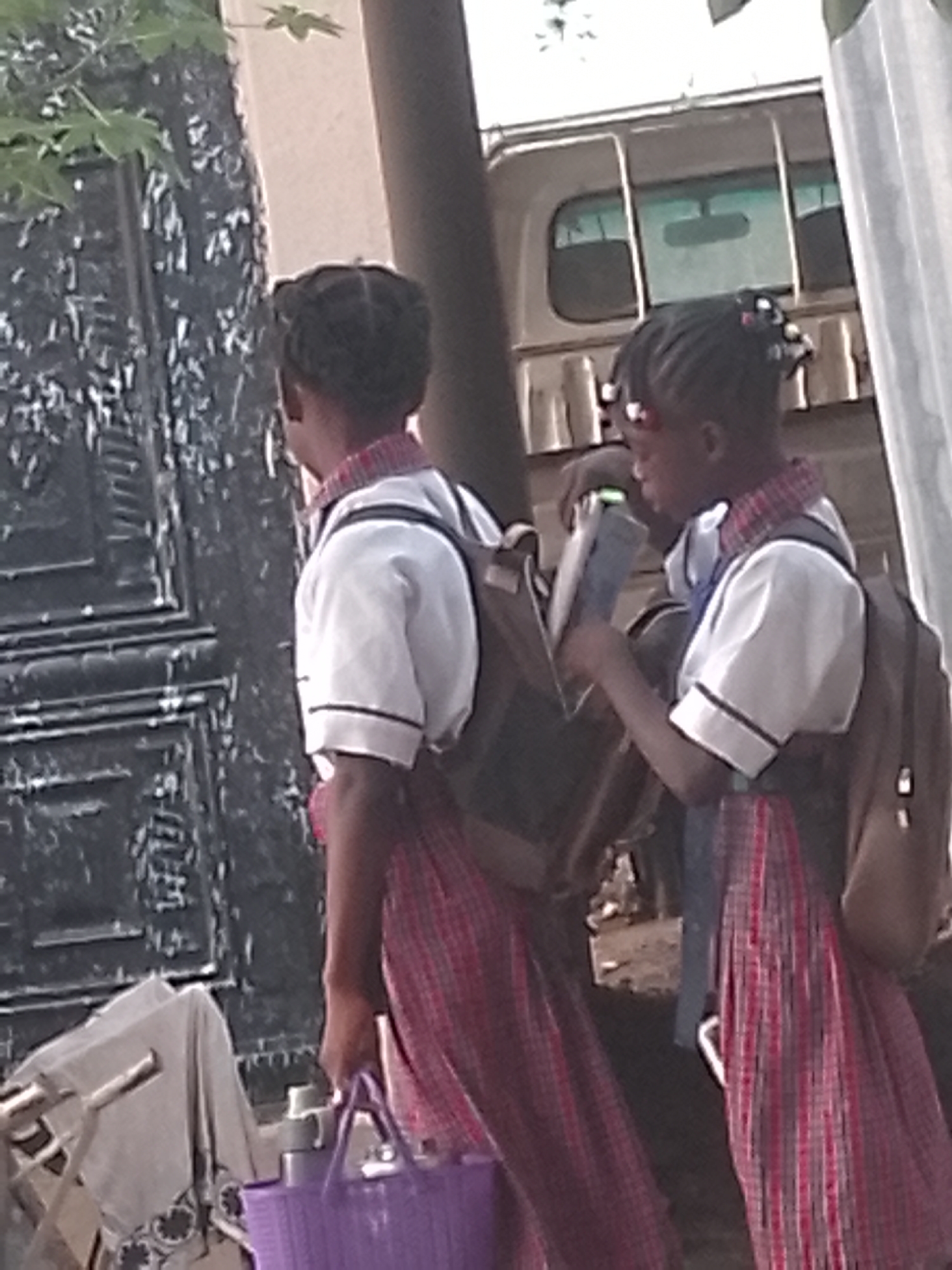 Two young girls in school uniforms stand on a city street in Unknown, Unknown. They wear white, short-sleeved shirts with black trim and red plaid skirts. Each carries a backpack. The girl on the right holds a book. The girl on the left holds a purple plastic basket. They stand in front of a dark, ornate door and a light-colored vehicle.