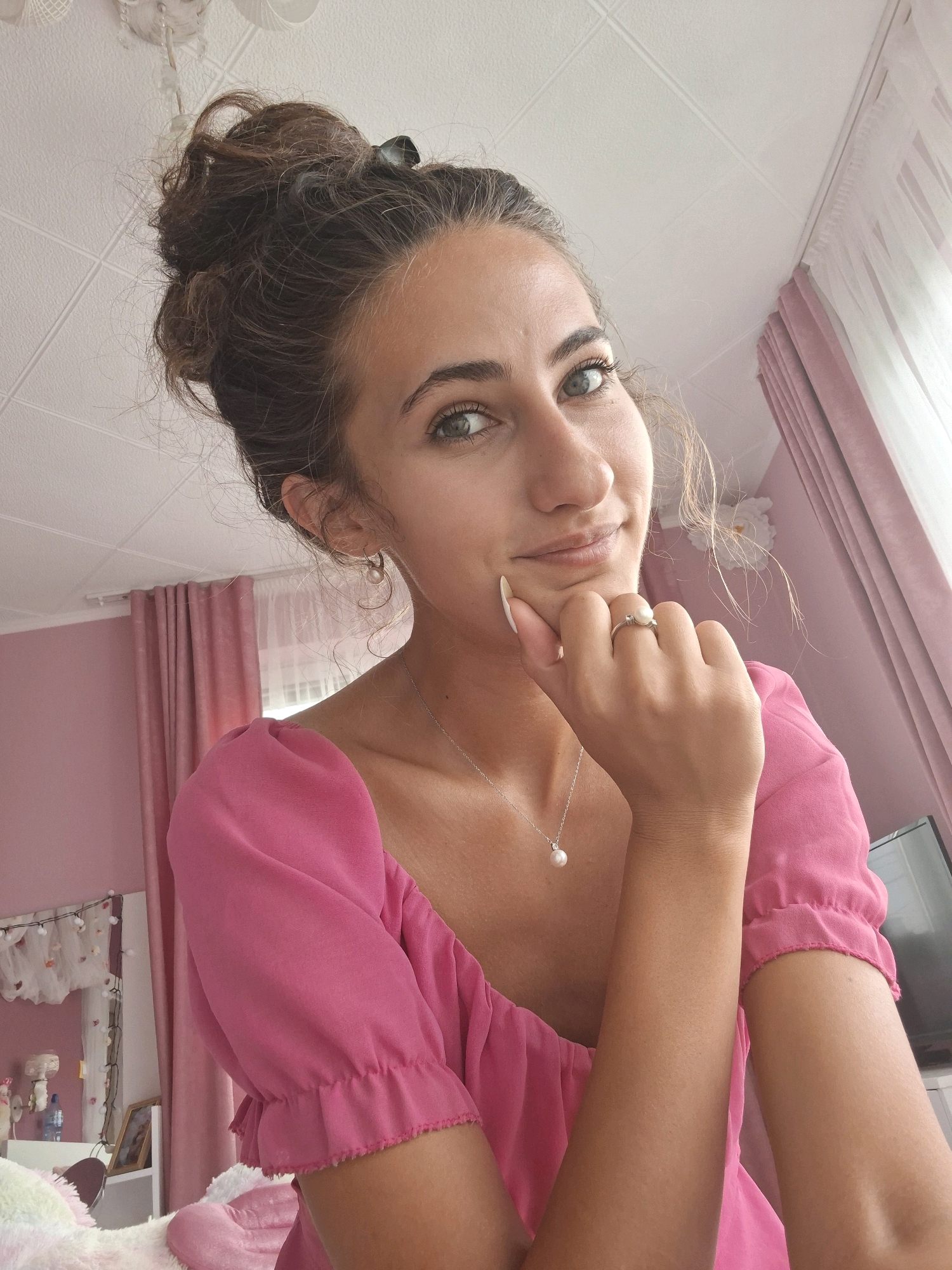 A young woman with dark brown hair pulled into a messy bun poses for a selfie in what appears to be a bedroom. She is wearing a pink, off-the-shoulder top with ruffled sleeves. She has a pearl necklace and a pearl ring on her left hand, which she is resting on her chin. Her eyes are green and she has a slight smile. The background is decorated with pink curtains, a white ceiling with a tiled pattern, and some lights strung along a mirror. The scene is well-lit and appears to be indoors.