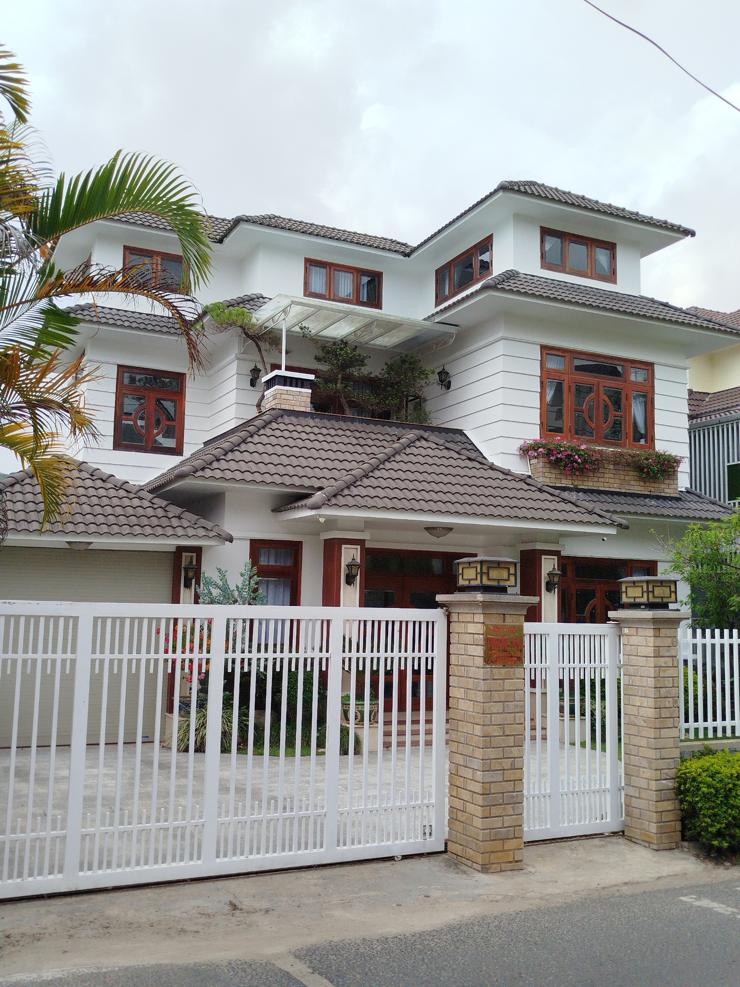 This full-shot image presents a large, multi-story house in a residential setting, likely in Da Lat, Vietnam, given the provided context. The house features a contemporary design with white walls and multiple brown, distinctively tiled roofs across its various levels.
The architecture includes numerous windows with warm, reddish-brown wooden frames, some of which exhibit ornate circular patterns. On the second floor, a prominent window boasts a flower box filled with vibrant pink and purple blossoms. An upper-level balcony or terrace is partially covered by a glass canopy, and green foliage is visible around it. A decorative brick chimney-like structure adds detail to the left side of the house.
In the foreground, a white metal gate with vertical bars secures the property. It comprises a wide section for vehicles and a narrower pedestrian gate on the right. These gates are supported by sturdy brick pillars, each topped with a decorative lantern-style light fixture. A concrete driveway is visible behind the gate, leading to a garage entrance on the left. The yard within the gate is lush with various plants and bushes, and a large palm tree partially frames the house on the left. An asphalt road runs along the immediate foreground.
The scene takes place during the daytime, under an overcast or cloudy sky, contributing to a muted, soft light. No people or specific activities are depicted, emphasizing the house's architectural features and its well-kept surroundings.
A small, rectangular red plaque with white text is affixed to the right gate pillar. Although the text is not clearly legible, it appears to be in Vietnamese, consistent with the location context of Da Lat. The overall impression is one of a spacious and carefully maintained modern home.