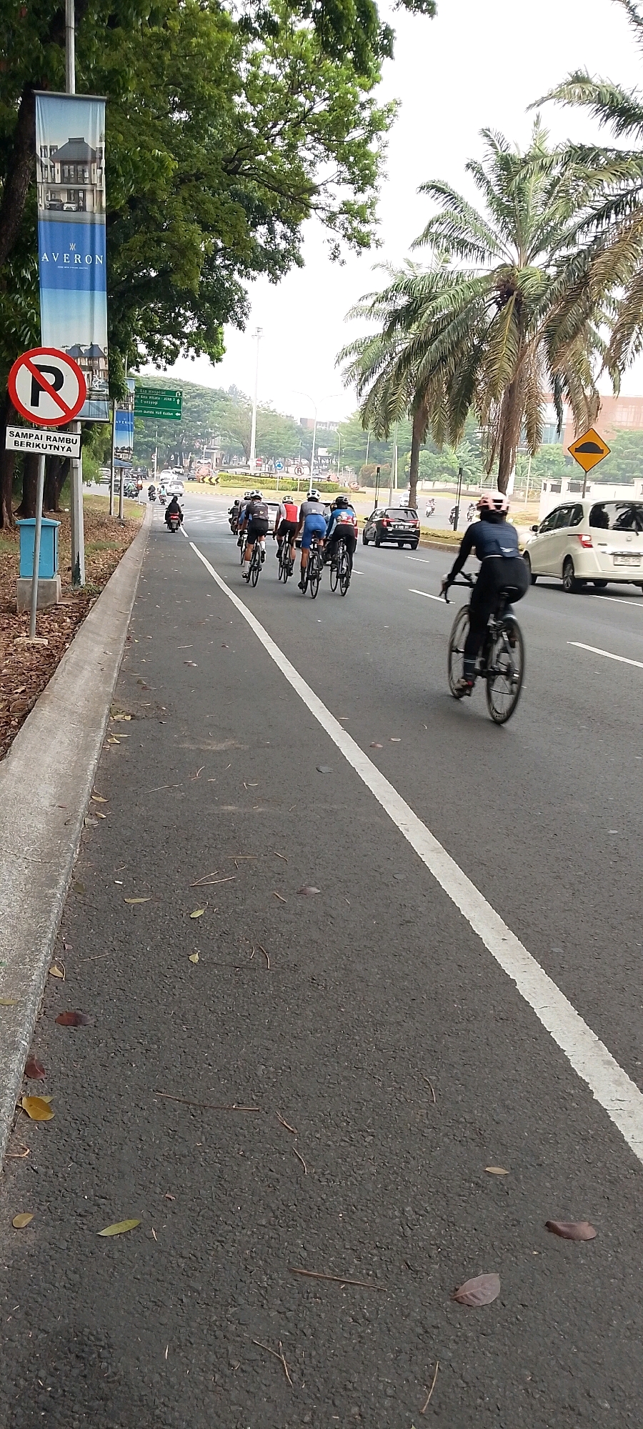 A group of cyclists rides on the right side of a paved road in Gunung Putri, Indonesia. The cyclists are in the middle of the road, and there is a lane to their right for other traffic. There are also pedestrians on the left side of the road, on the sidewalk. The road is lined with trees, and there are buildings in the background. The sky is overcast. A banner with the text "AVÉRON" hangs from a lamppost on the left side of the image. There is also a red and white "no parking" sign. The road has a white line marking the lane. Several cars and a motorcycle are visible on the road.