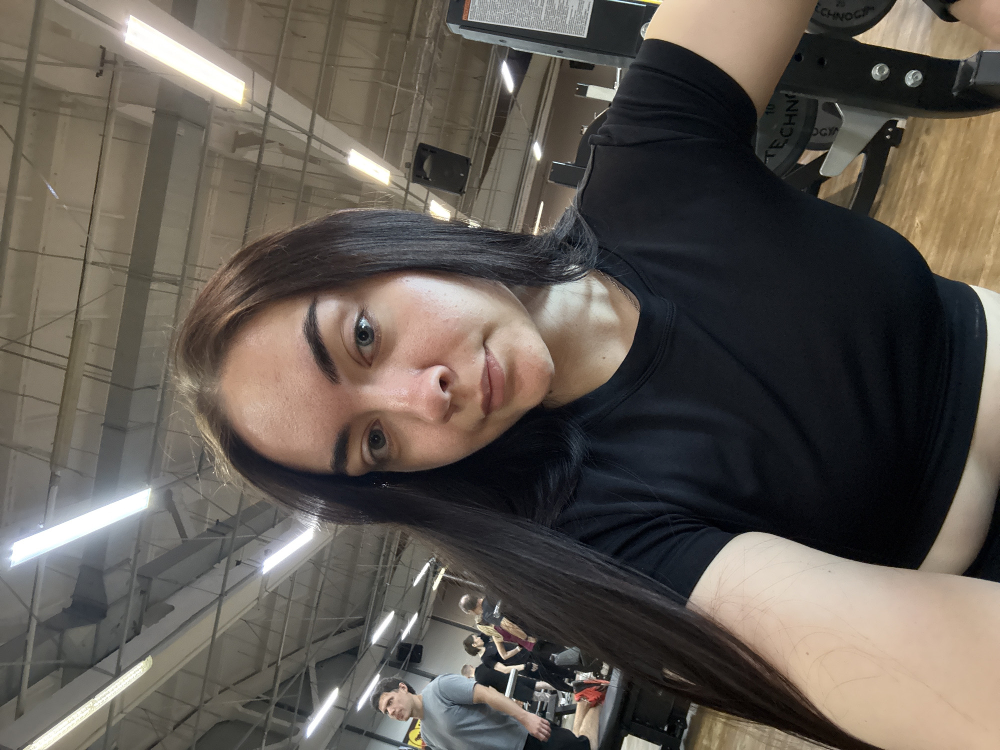 A selfie of a woman with long brown hair wearing a black t-shirt in a gym, presumably in Chaikovsky, Russia. Other individuals and exercise equipment are visible in the background. The overhead lighting is fluorescent.