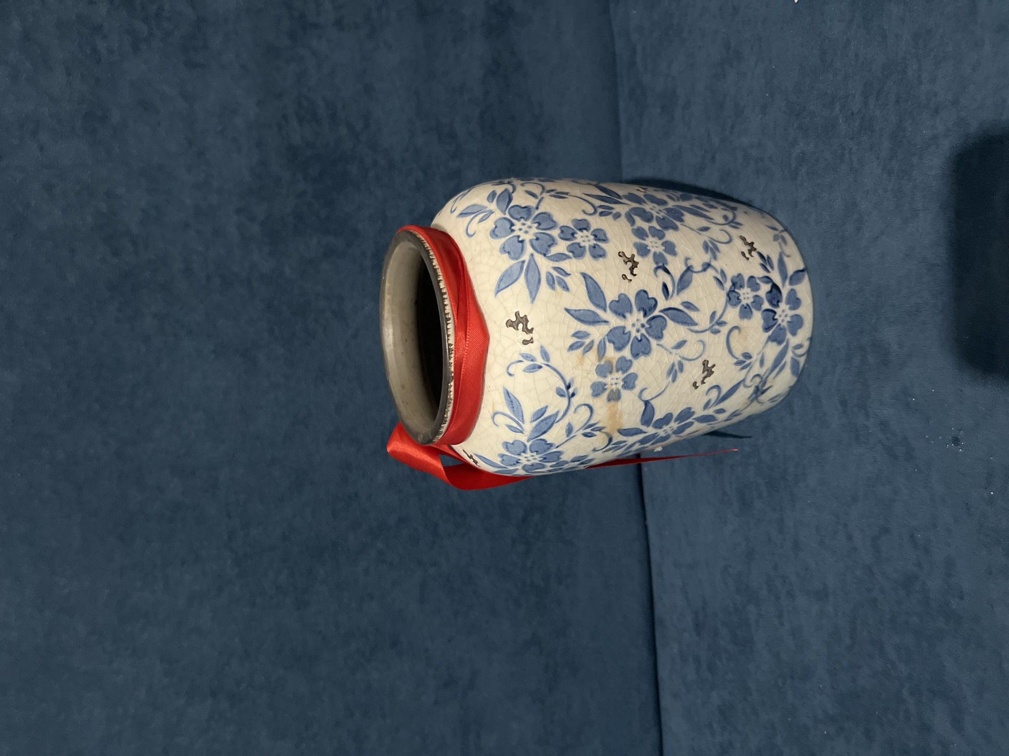 A ceramic vase with a blue floral pattern and a red ribbon tied around its neck sits on a dark blue textured surface. The vase is positioned slightly off-center to the right, with a portion of its body and opening visible. The background is a solid dark blue fabric, likely a sofa or backdrop, with subtle shadowing and texture. There are no people or any indication of the location being within the city of Bishkek City, Kyrgyzstan.