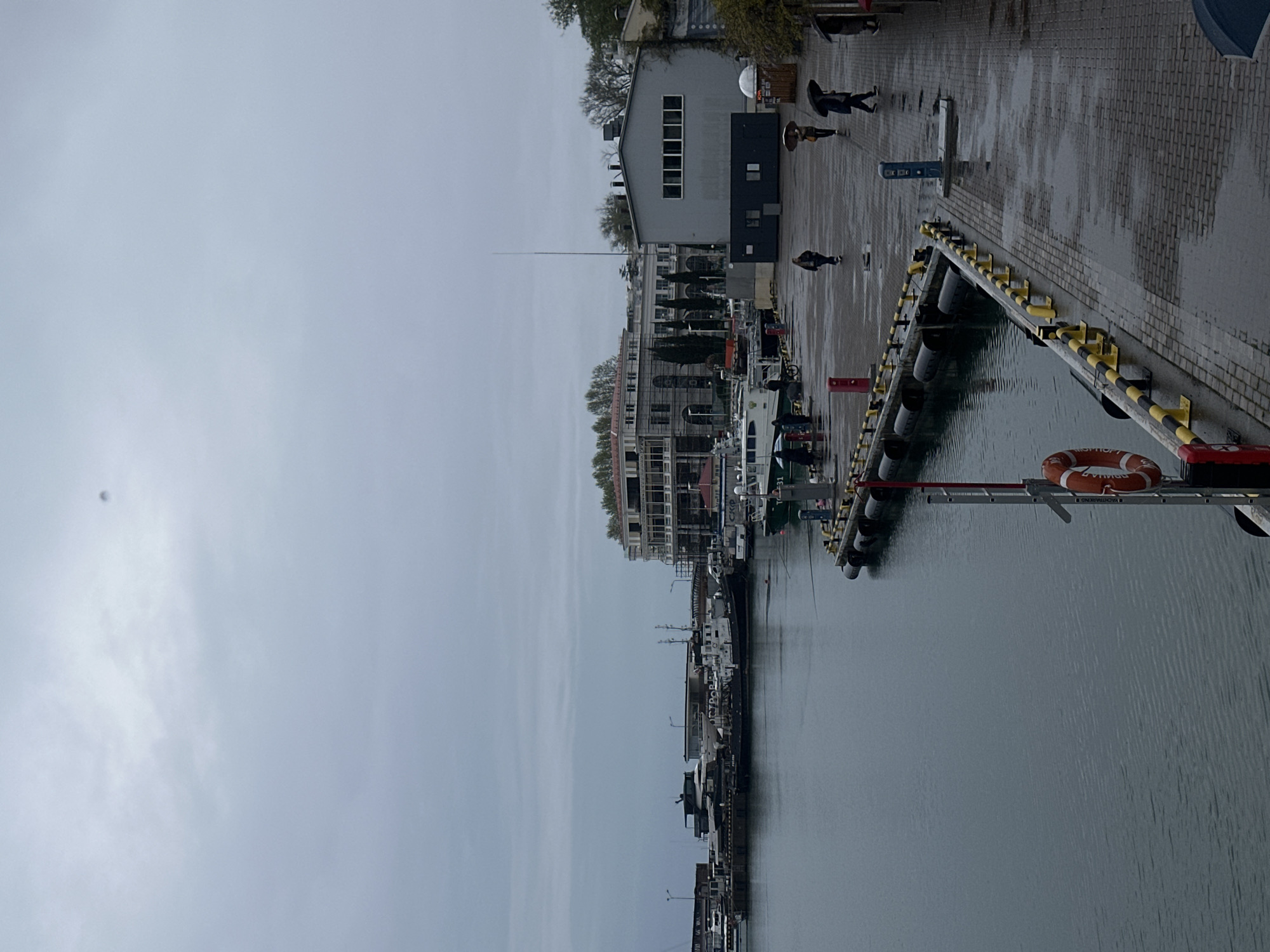 The image shows a harbor in Sochi, Russia, on an overcast day. Several boats are docked along a pier. A grey building stands at the edge of the pier, adjacent to a pedestrian walkway where people are walking. A life preserver hangs on the edge of the pier. The sea is calm.