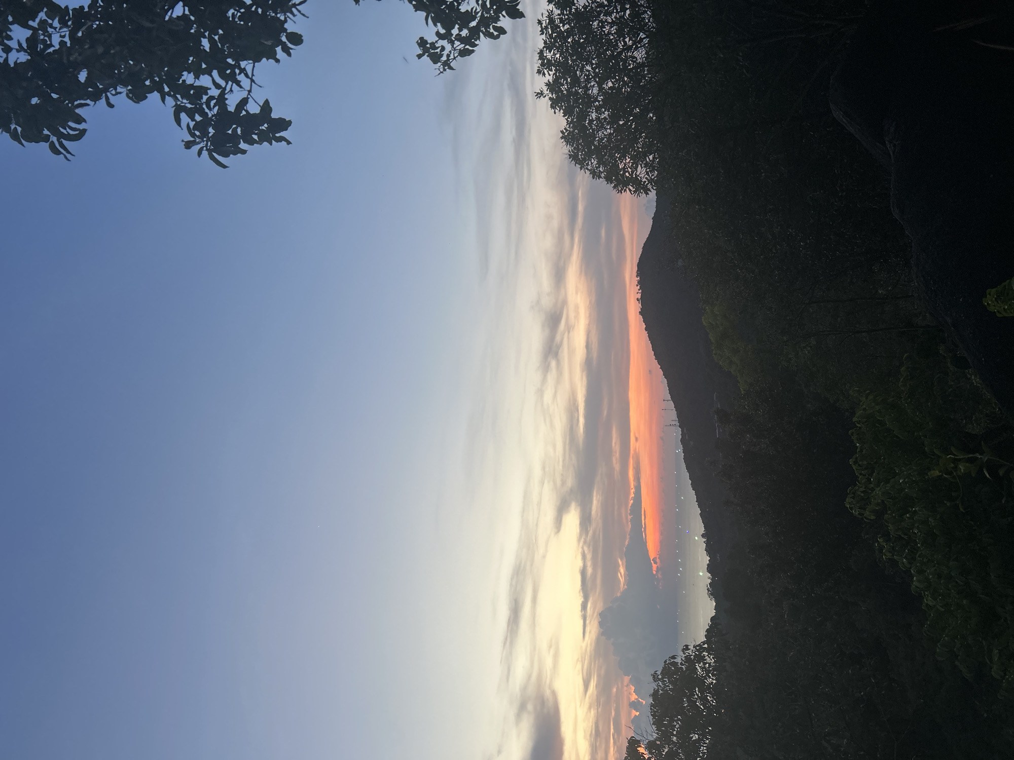 The image shows a view from Baan Hin Kong, Thailand, during a sunset or sunrise. The sky has colors ranging from blue to orange and pink, with light clouds. Dark tree foliage is visible in the foreground along the top and right side of the image. In the distance is a dark mountain ridge with what appear to be lights on the slope.