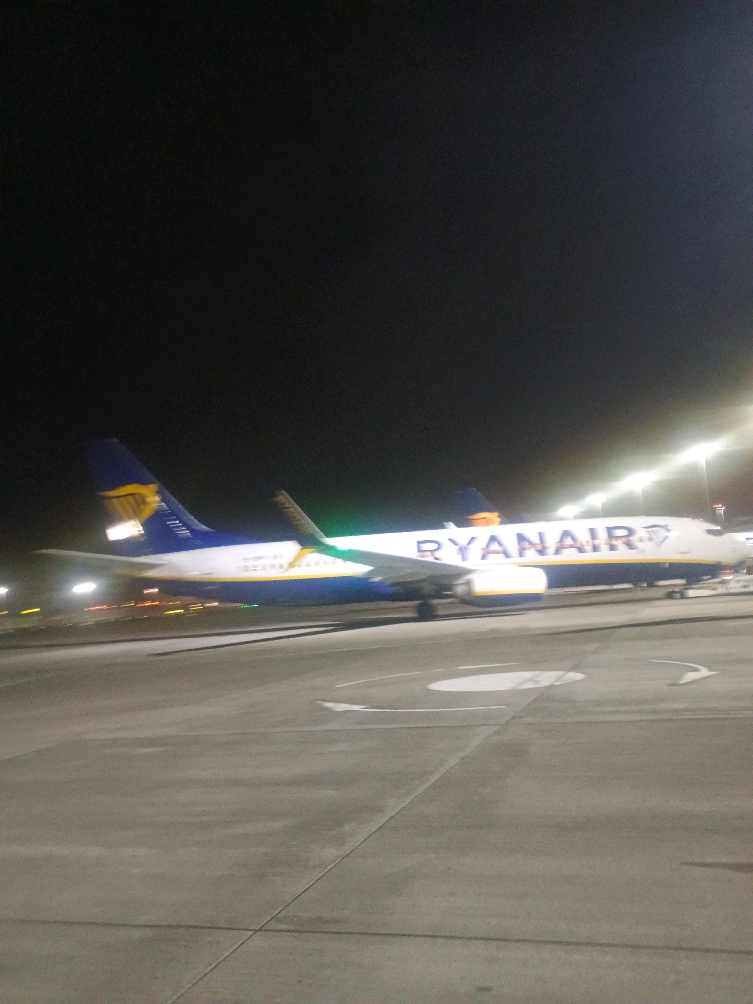 An undated, vertical, low-light photograph captures a side view of a Ryanair airplane parked on an airport tarmac at night. The airplane, predominantly white with blue and yellow accents and the distinctive "RYANAIR" logo emblazoned on its fuselage, is the central subject. Its wings and tail are visible, with the tail fin featuring the Ryanair logo and yellow stripes. The tarmac is concrete, marked with white lines and a circular symbol, indicating it's an operational area. In the background, tall airport lighting poles cast a bright glow, illuminating the scene and creating streaks of light due to the low light conditions and potential movement. The night sky is dark, devoid of stars or moon. The image is slightly blurred, suggesting it was taken from a moving vehicle or with a handheld camera in low light. No people are visible in the frame.
