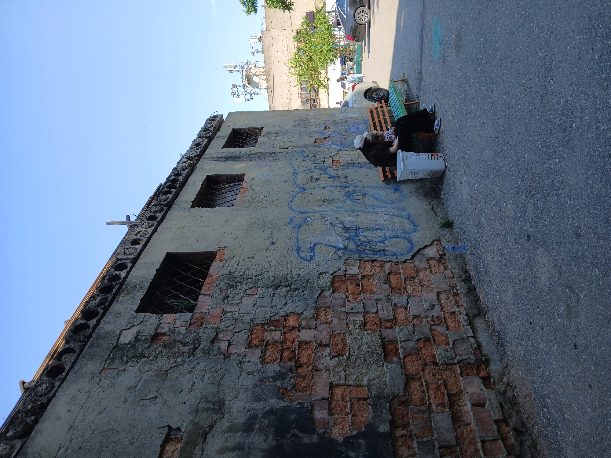 A woman wearing a white hat and dark clothing is sitting on a green and orange park bench next to a child. The woman appears to be holding a phone. A white trash can is located to the left of the bench. The bench and trash can are situated on an asphalt surface next to a damaged concrete wall covered in graffiti. Behind the wall, there are buildings and communication towers visible. A car is parked to the right of the bench, and another car is driving in the background. The sky is clear and blue. The image appears to have been taken in Makhachkala, Russia, based on the architectural style and the presence of communication towers typical of the region.