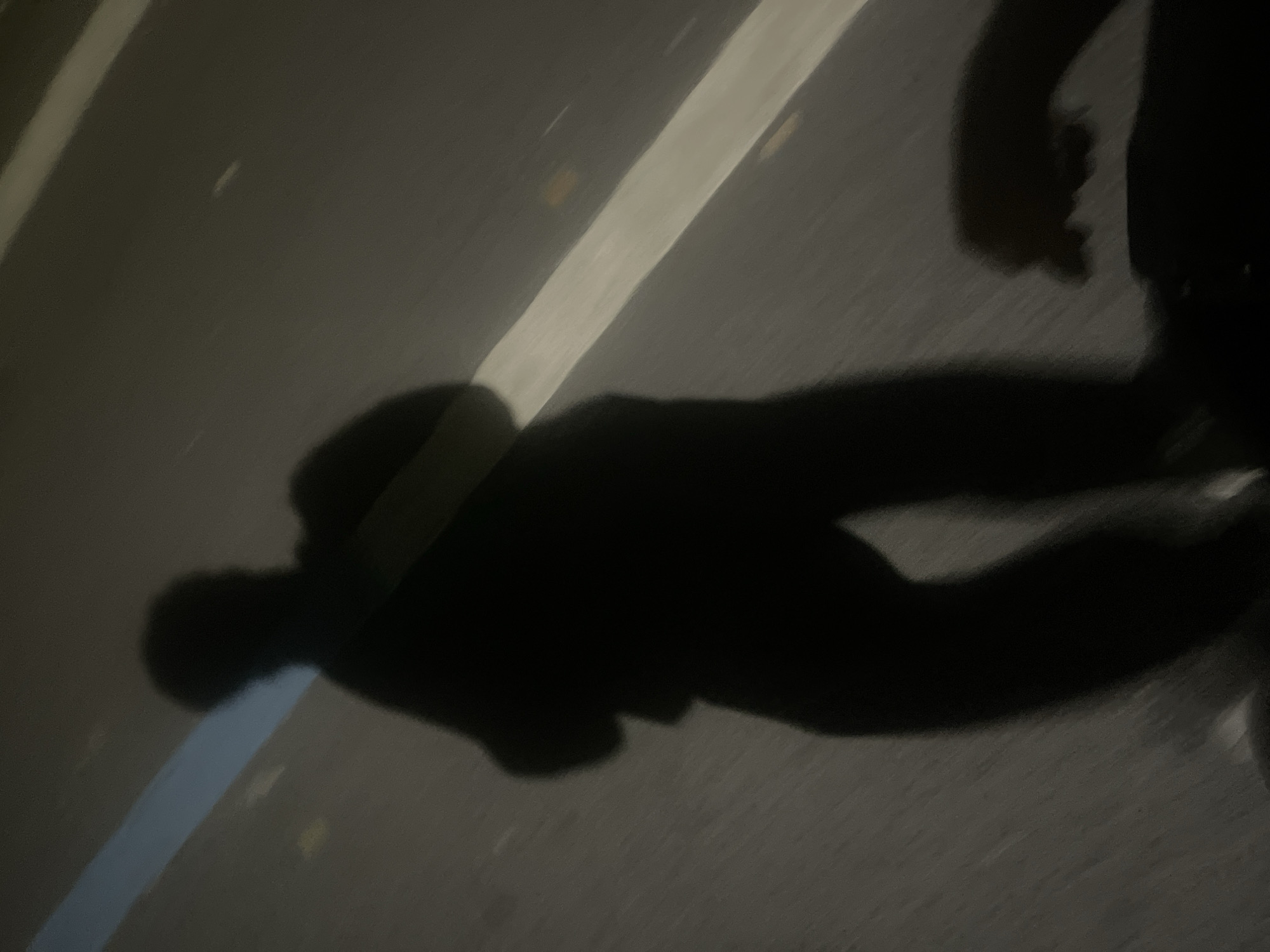 The image appears to be taken at night on a road or street, possibly in the Chongqing High-tech Zone, China, given the prompt. The primary subject is a shadow cast by a person wearing what appears to be a backpack or carrying something on their back. The shadow also shows the person's arms and legs in motion, suggesting walking or running. The surface is likely asphalt, with lane markings visible as light-colored lines. The overall environment is dark, with limited lighting.