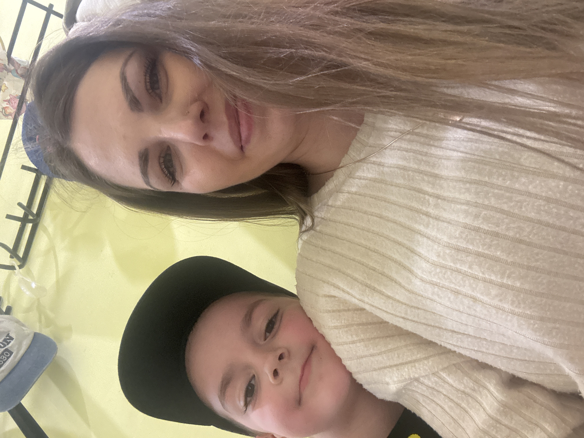 A selfie shows a woman and a child. The woman has long brown hair, fair skin, and dark eyelashes. She wears a light-colored sweater. The child, wearing a black baseball cap, is next to her. The background is light yellow. A hat rack with a blue baseball cap is visible in the lower-left corner. The location in Shevlyakovo, Russia cannot be confirmed.
