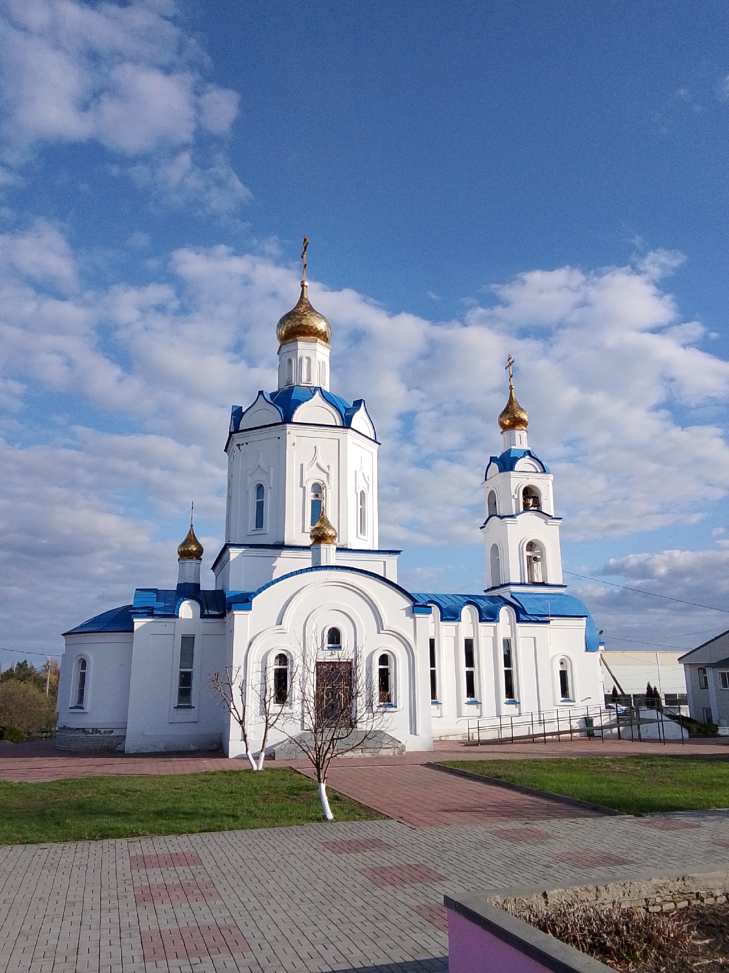 The image displays a beautiful Orthodox church, located in Khlevnoye, Russia. The church is predominantly white with contrasting bright blue roofs and multiple gleaming golden domes, each crowned with a golden Orthodox cross. The architecture features a large central dome, several smaller domes, and a prominent bell tower on the right side, which also has a golden dome and arched openings revealing bells within.
The scene is set under a clear, vibrant blue sky adorned with scattered white, fluffy clouds, indicating good weather, likely late morning or early afternoon due to the bright and even illumination. In front of the church, there is a neatly kept green lawn bordered by a paved pathway made of red and grey bricks that leads towards the main entrance. Two small, bare trees with whitewashed lower trunks stand symmetrically near the church's entrance.
In the background, on the left, distant green foliage from trees is visible. To the right of the church, parts of other buildings can be seen, including a grey structure and a larger, possibly industrial, building further back. A wheelchair ramp with a dark railing provides accessibility to the church's side entrance. No people are present, and no specific activity is taking place. There is no visible text in the image.