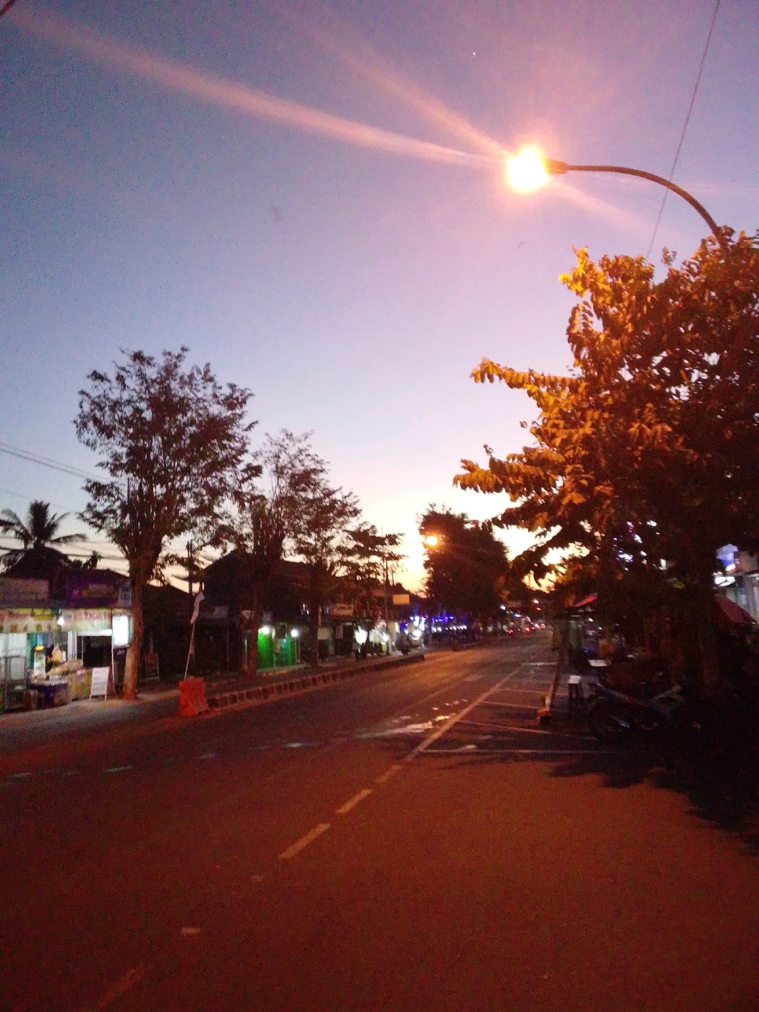 The image captures a street scene in Purworejo, Indonesia, during twilight. The sky exhibits a gradient of purple and pink hues, with a bright streetlamp illuminating the right side of the frame. Several trees with dense foliage are visible along the roadside. On the left, a row of shops with illuminated signs and a few people are present. The street itself is paved with asphalt, marked with dashed white lines. A few motorcycles are parked on the sidewalk to the right. The overall atmosphere suggests a bustling street at the end of the day.