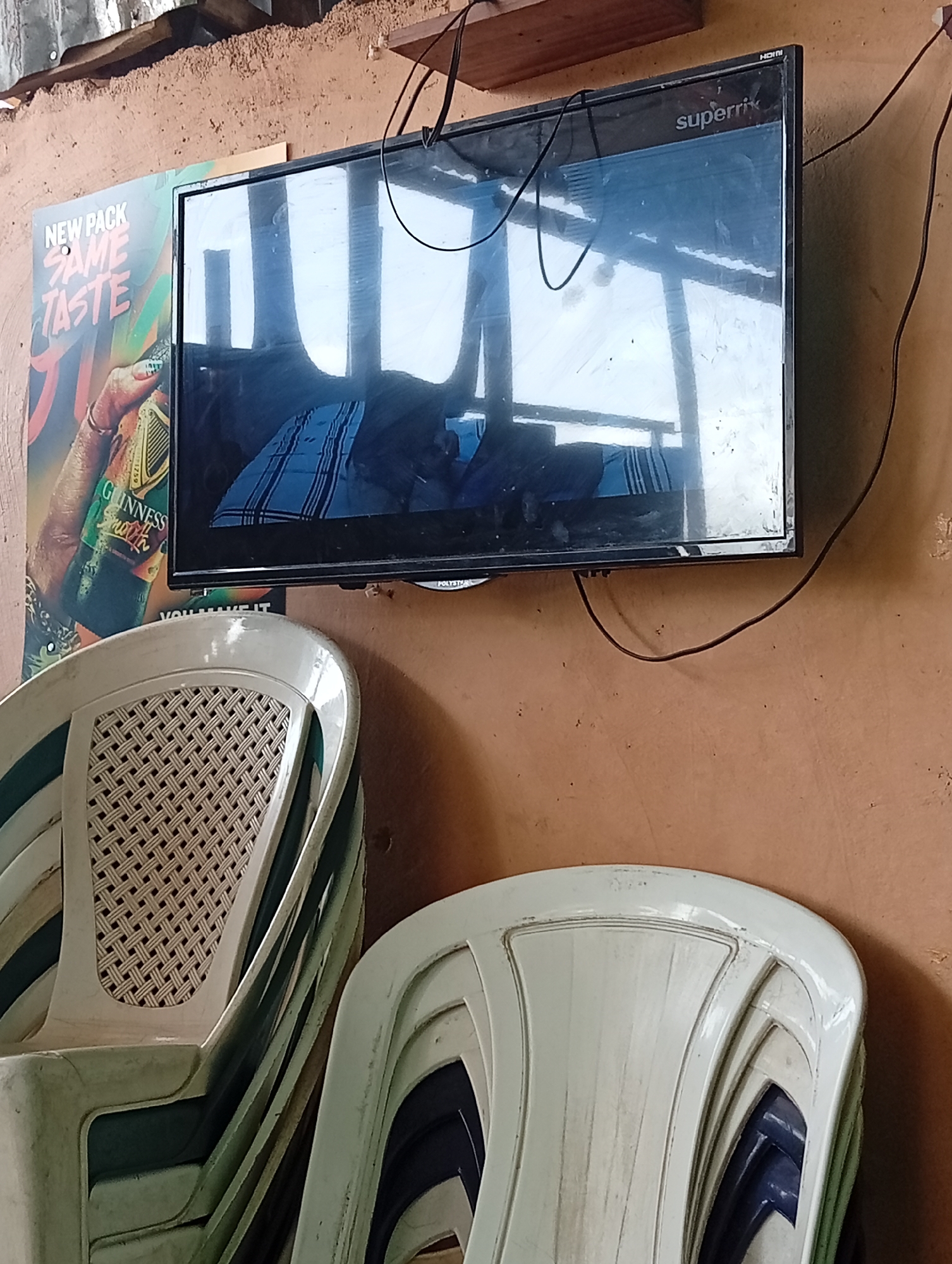 The image shows a stack of white plastic chairs beneath a "superrik" brand television mounted on a brown wall. To the left of the television is a poster advertising Guinness stout with the words "NEW PACK SAME TASTE". The wall appears textured. The reflection on the television screen obscures the image that is playing, but it is reflecting an open space with corrugated roofing. The environment suggests a casual, possibly commercial setting such as a small bar or restaurant. The image was captured in Jos, Nigeria.