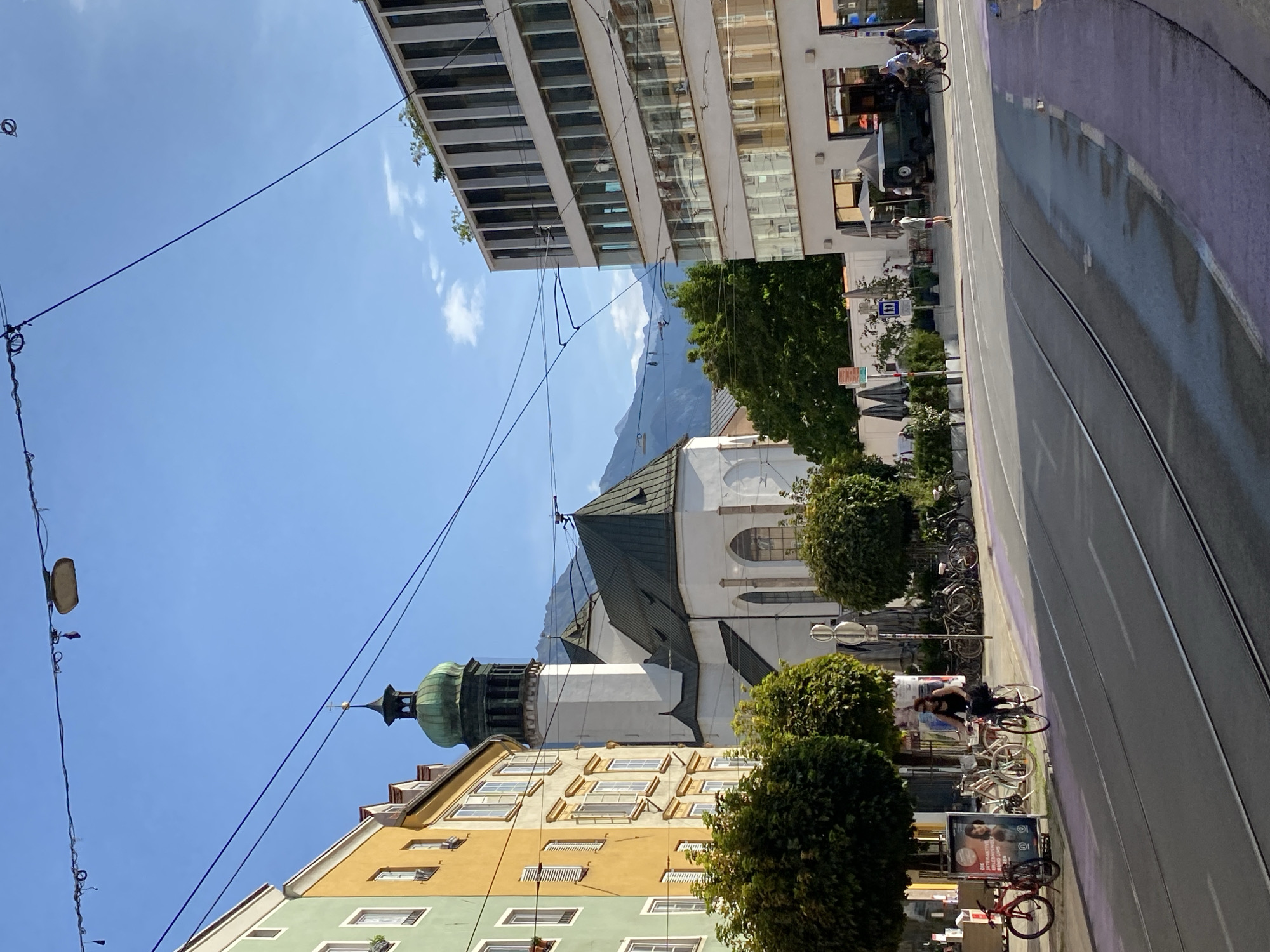 The image captures a street scene in Innsbruck, Austria, with a view of the Innsbruck Cathedral (Hofkirche) and its distinctive green dome in the background. The foreground shows a tram line running along the road, with a purple-painted bike lane adjacent to it.
In the middle ground, a woman is riding a bicycle in the bike lane. Several other bicycles are parked along the sidewalk. A green jeep is parked on the side of the road near a cafe with outdoor seating.
The architecture of the buildings is varied, with some older-style buildings on the left painted in yellow and green, and a more modern building with a glass facade on the right. Trees line the street, and mountains are visible in the far background under a clear blue sky. The scene appears to be from a sunny day, with shadows cast by the buildings and trees.