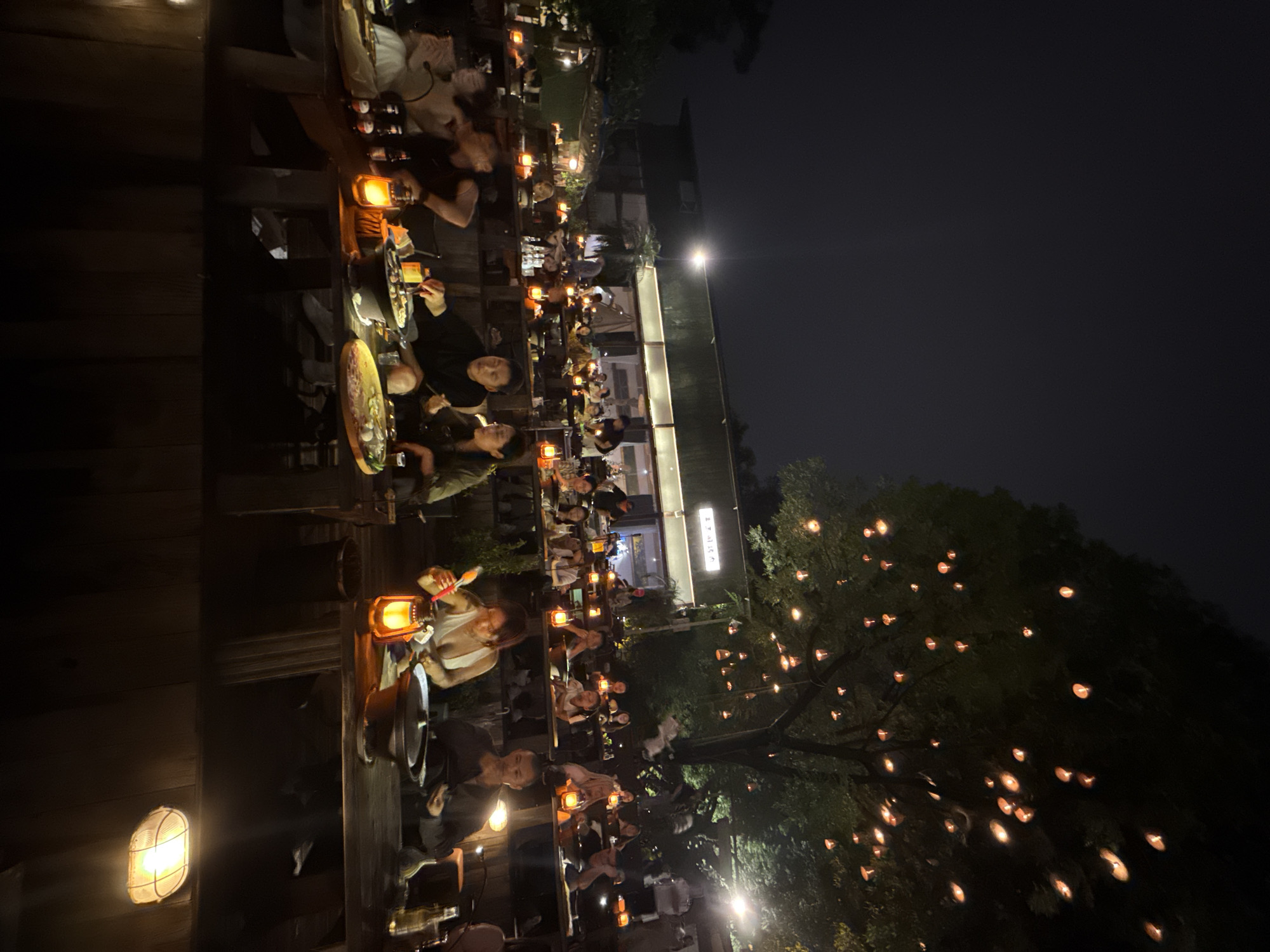 The image depicts an outdoor dining area at night, likely a restaurant or gathering place, in Nan'an District, Chongqing, China. Multiple long tables are arranged linearly, occupied by patrons who appear to be dining and socializing. Each table is lit by individual lanterns or lamps, creating a warm, ambient light. In the background, a building with illuminated signage is visible, suggesting a commercial establishment. To the right, a large tree is adorned with numerous small lights, contributing to the festive atmosphere. The overall setting suggests an evening event or a lively restaurant scene. The image orientation is vertical.