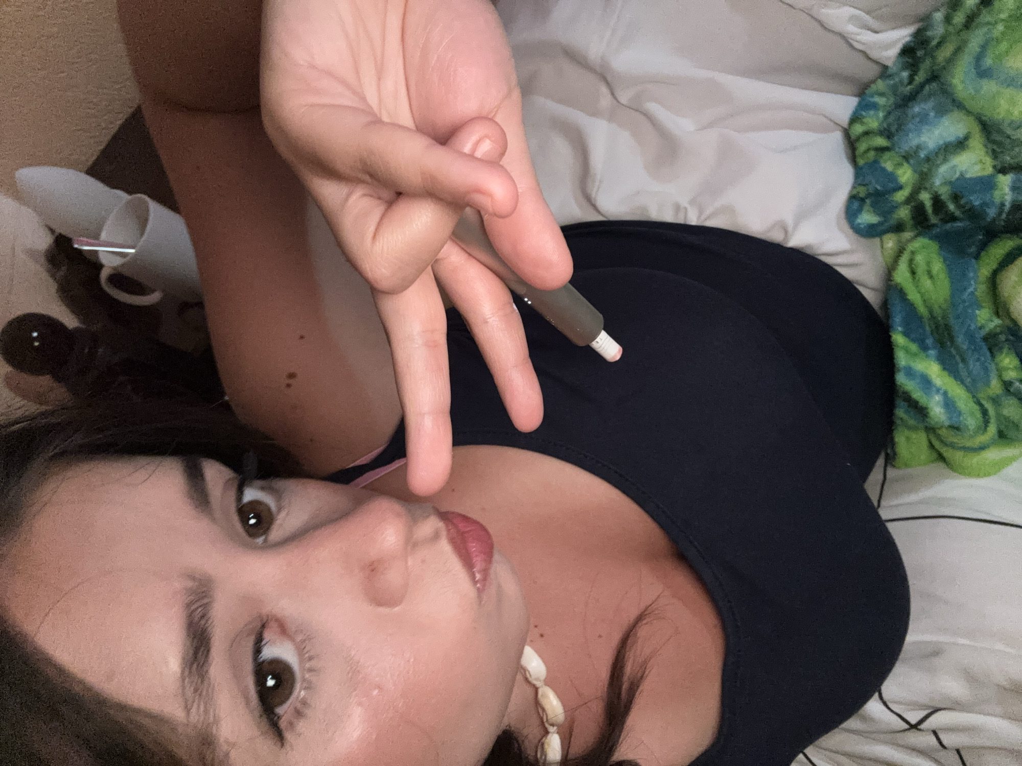 A close-up shot shows a woman lying on a bed, holding a vape pen in her right hand. She is wearing a navy blue tank top and a shell necklace. Her hair is dark and her eyes are brown. The background is a white bedsheet with a crumpled green and blue patterned blanket in the lower right corner. The image was taken in Garmen, Bulgaria.