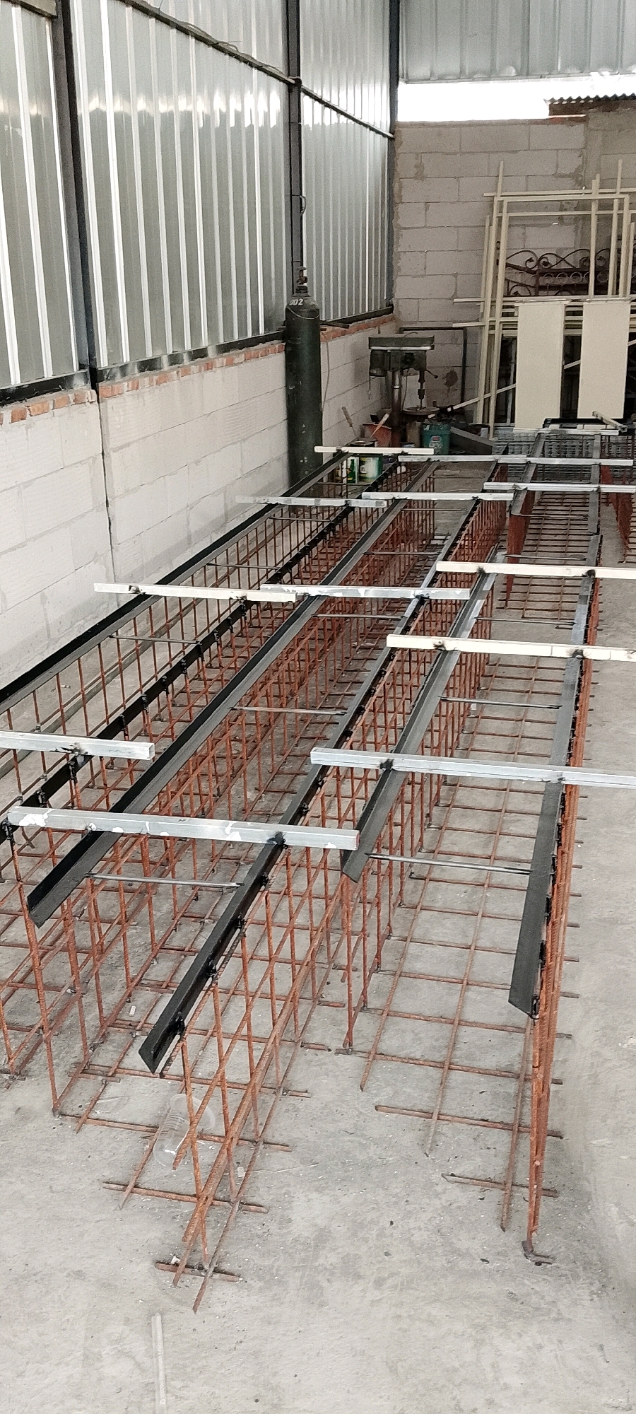 An indoor construction site in Sriamur, Indonesia features rows of rebar cages laid out on a concrete floor. The cages are rectangular and appear to be awaiting concrete pouring. Metal beams are positioned horizontally across the top of some cages, secured with clamps. In the background, cinder block walls and a corrugated metal roof are visible. A green gas cylinder stands near the wall. Metal framing and white panels are stacked in the far background, suggesting ongoing construction or fabrication work. The scene is illuminated by natural light entering from an opening in the roof.
