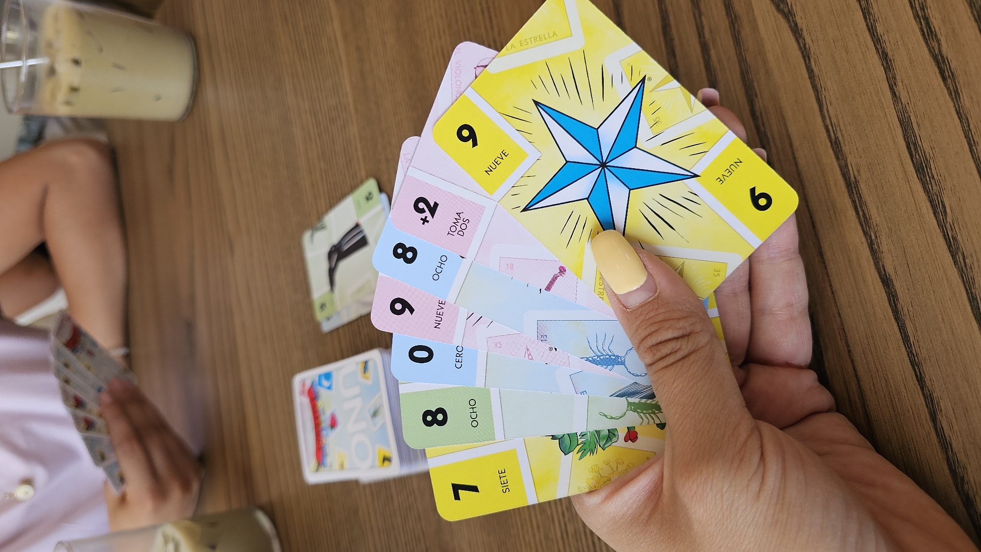A medium, eye-level shot displays a hand holding a fan of lottery-style cards over a striated, wood-grain surface, near a fair-skinned individual. The surface, likely a table, has a partly visible glass containing a light-colored beverage on the upper left.
The cards, fanned out in the hand, are printed with bold numbers, icons, and text in both English and Spanish. The top card features a yellow background with radiating lines and a blue star, with the number "9" at the upper left and lower right corners. Other cards in view display the numbers 7, 8, 0, 9, and "+2", along with related text in Spanish such as "OCHO," "NUEVE," "SIETE," "TOMA DOS," and "CERCO." One card has the word "UNO" visible on it. The subject’s fingernails are painted a pale yellow.
The person on the left is only partially visible, sitting behind the table, holding cards in their hand.
The location, San Bernardino Tlaxcalancingo, Mexico, is not directly discernible from the image.
