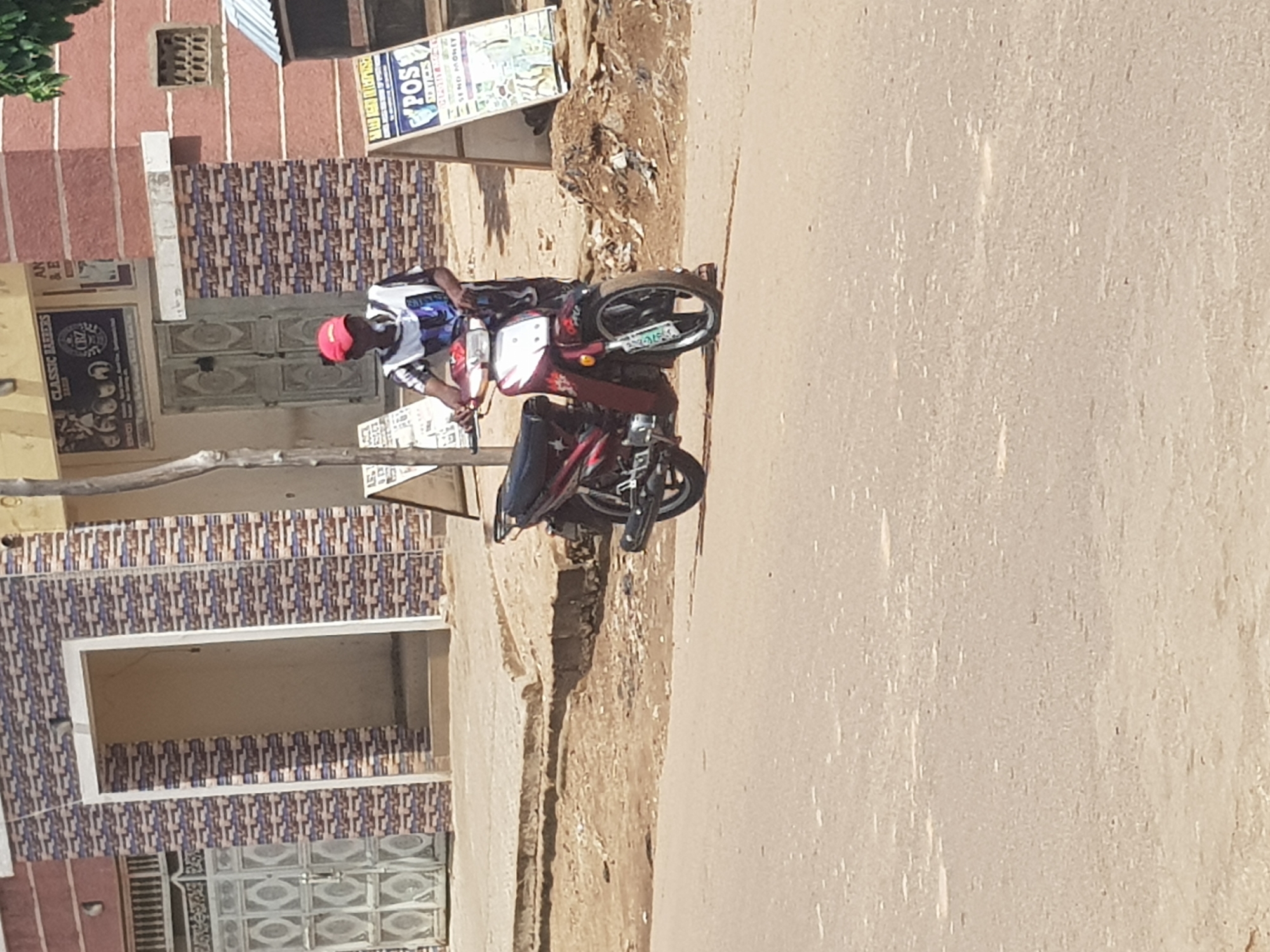 In Kufena, Nigeria, a person wearing a red cap and a striped t-shirt stands beside a red and black motorcycle on a dirt road. The motorcycle is parked on its kickstand. Behind the person and the motorcycle is a building with a patterned tile facade and an open doorway. To the right of the building, a sign with text and images is visible, and further to the right, another building with a red brick facade and a small, barred window is present. The ground is a mix of dirt and asphalt.