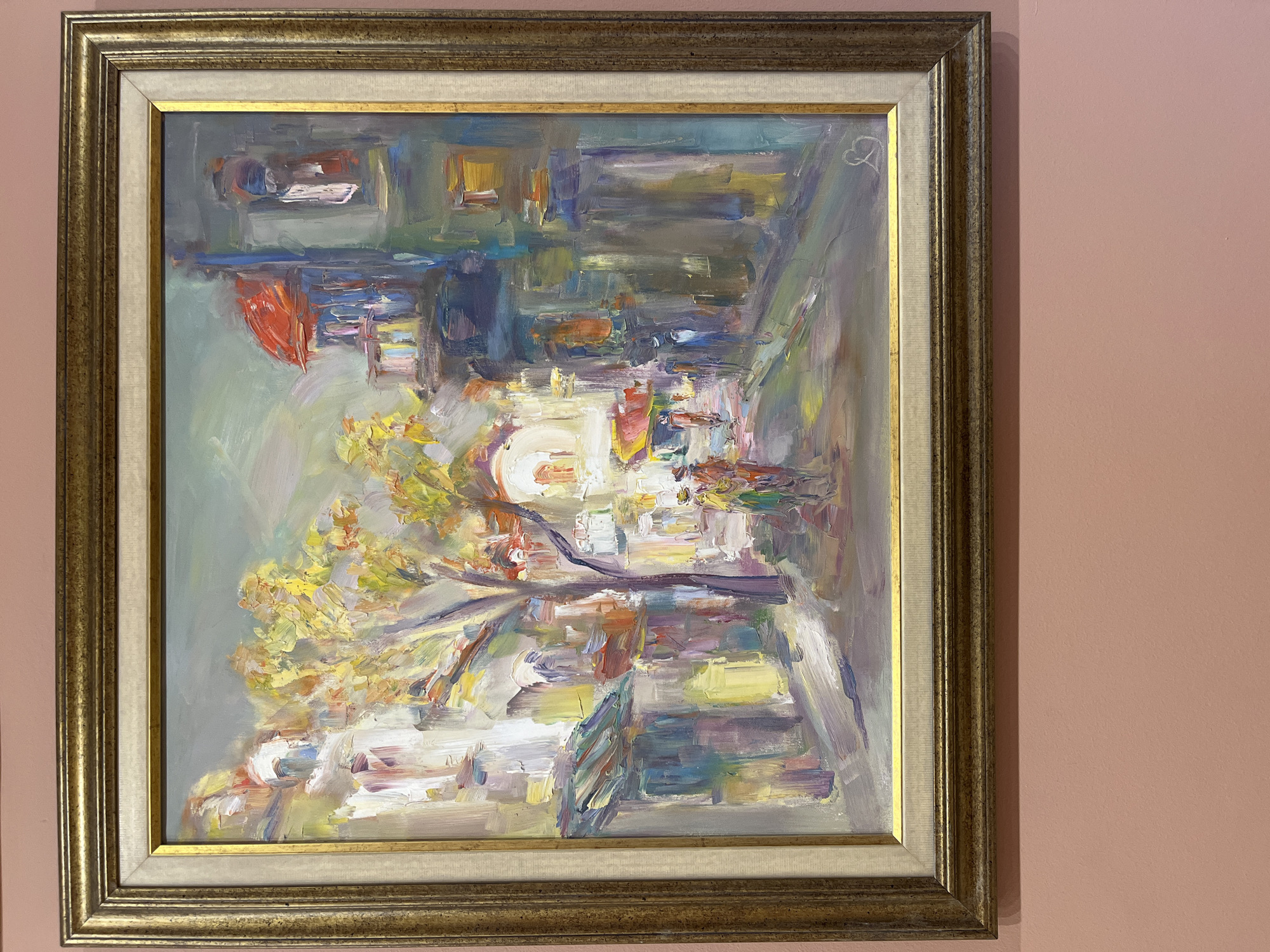 A framed oil painting hangs on a pink wall. The painting depicts an impressionistic scene, possibly of a street or square in a city. Yellow and white brushstrokes suggest foliage or blossoms, while other colors represent buildings, possibly in the old town of Plovdiv, Bulgaria. There are no clear depictions of people. The artwork has a signature in the upper right corner. The frame is ornate with a gold finish.